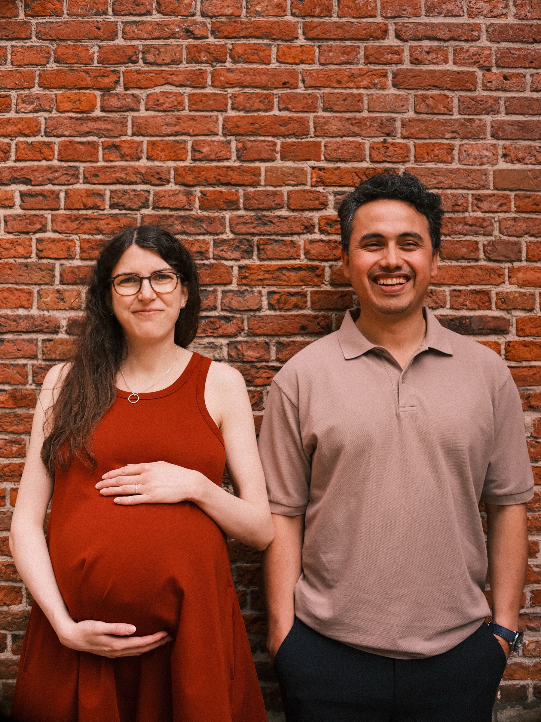 Expecting couple laughing in front of an old red brick wall in Leiden, her hands on her baby bump in a rust-red dress