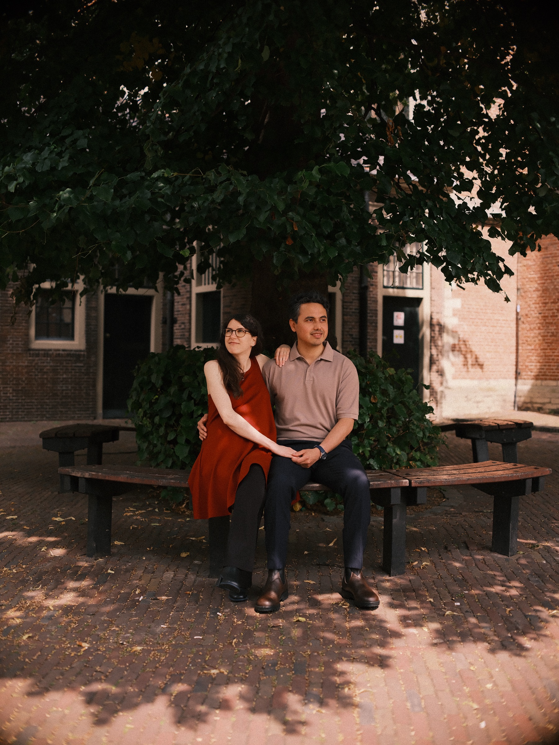 Expecting couple sitting close on a round wooden bench in dappled shade, her rust-red dress bright against the small brick houses behind