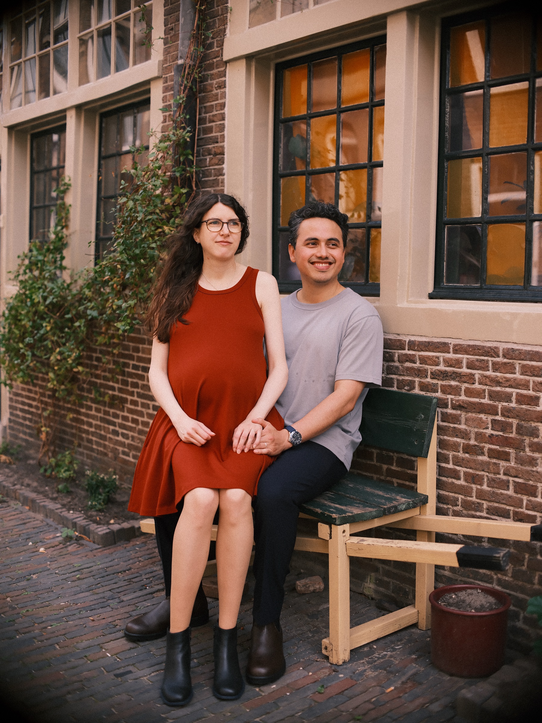 Expecting couple laughing on a wooden bench under lit windows, her hands resting on her baby bump and his arm around her