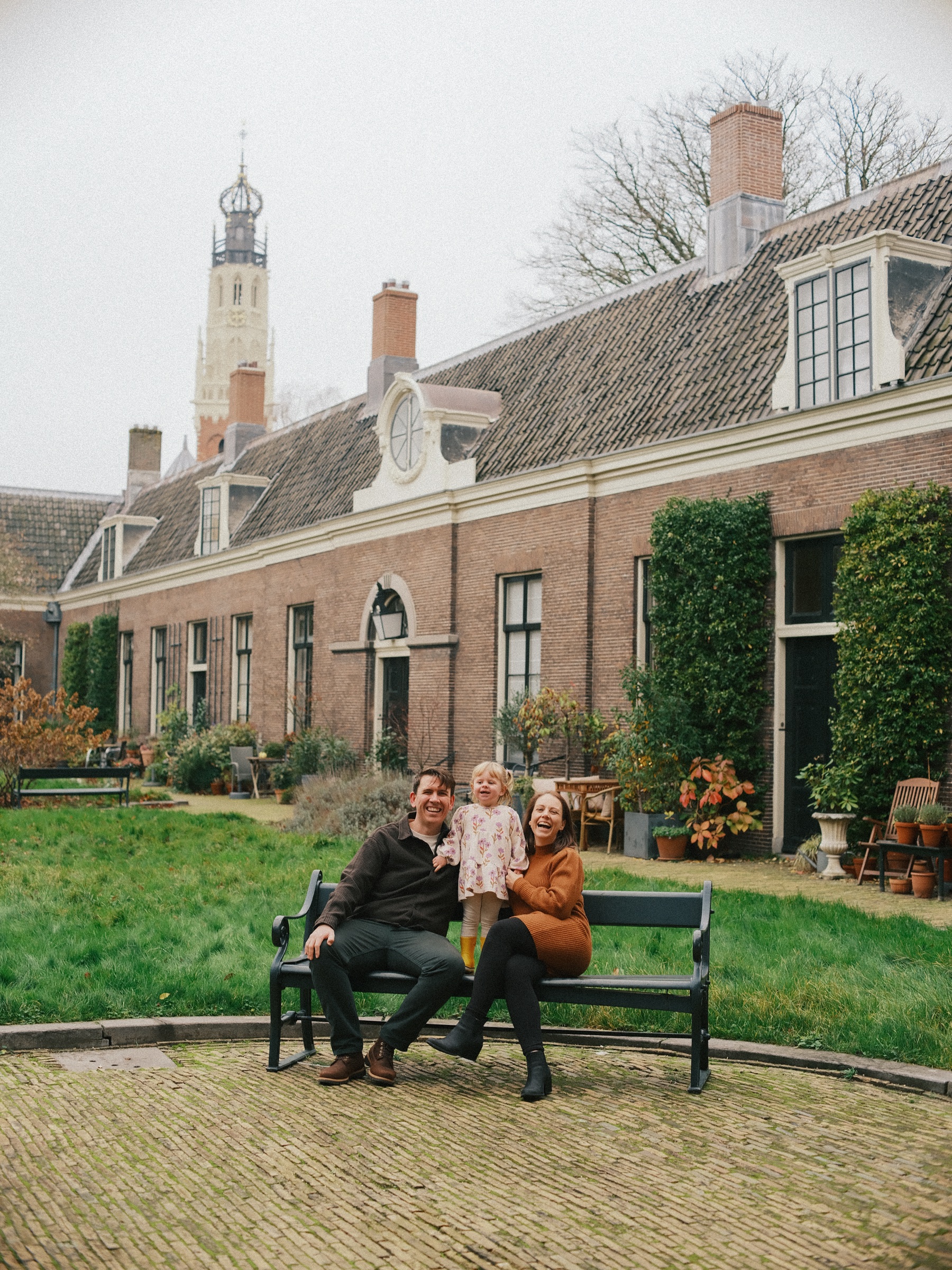Gezin op een groene bank in Teylers Hofje in Haarlem, met bakstenen huisjes, klimop en de witte toren van de Bakenesserkerk boven de daken
