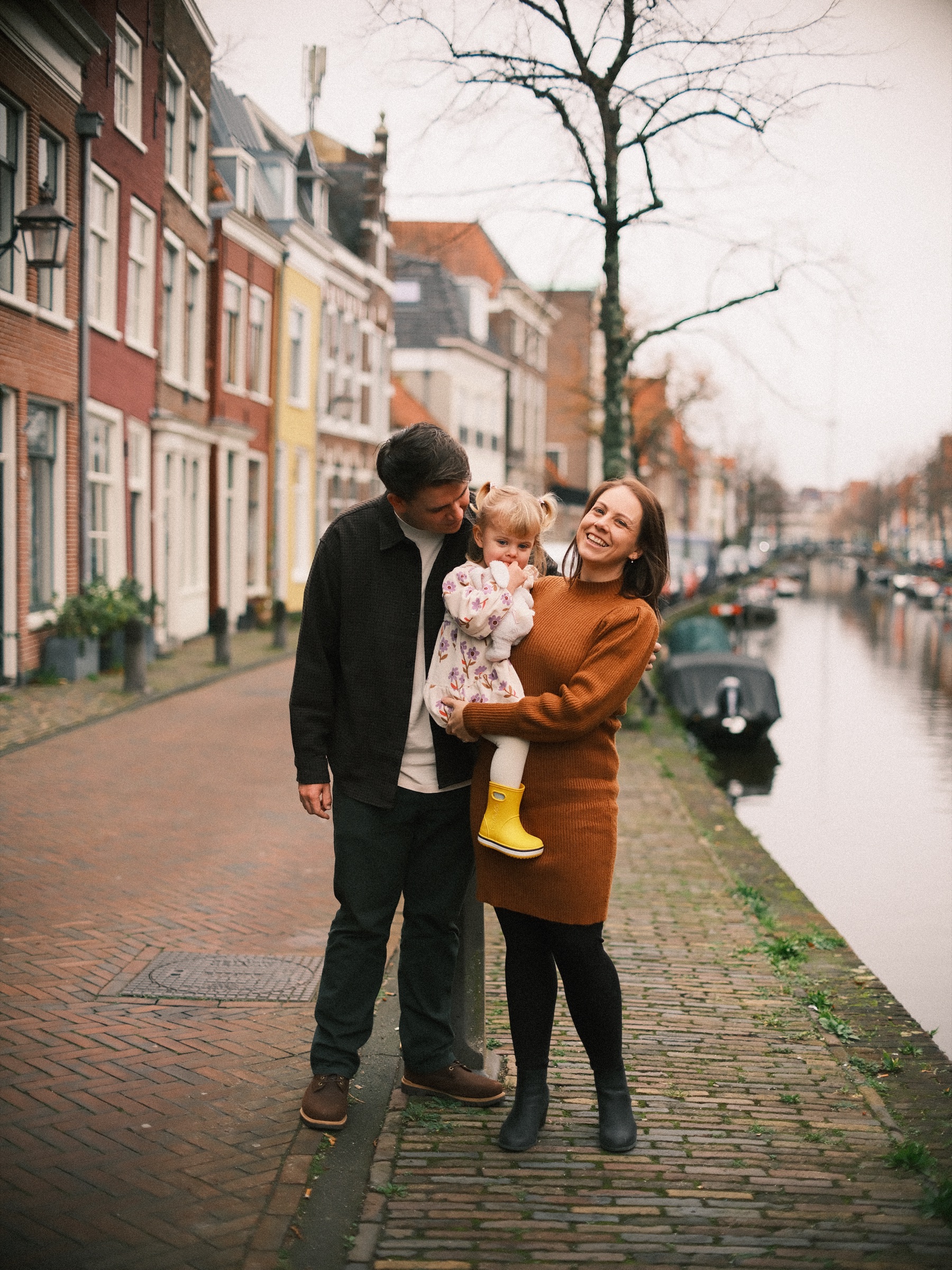 Moeder met hun peuter op gele regenlaarzen op de arm, de vader buigt naar ze toe, aan een gracht met puntgevels in Haarlem in november