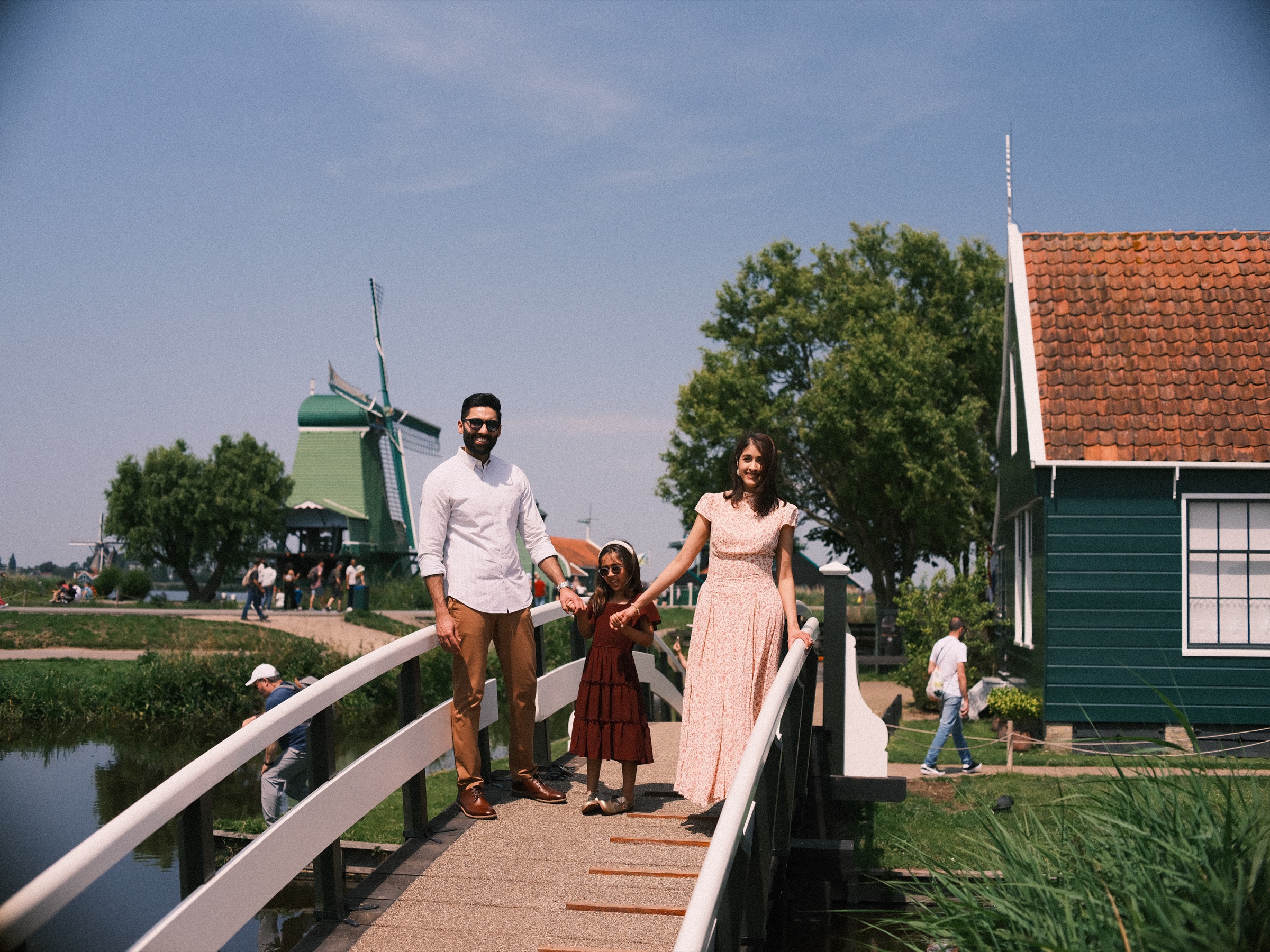 Family crossing a small white wooden bridge between green houses, a windmill behind them on their family trip to the Zaanse Schans
