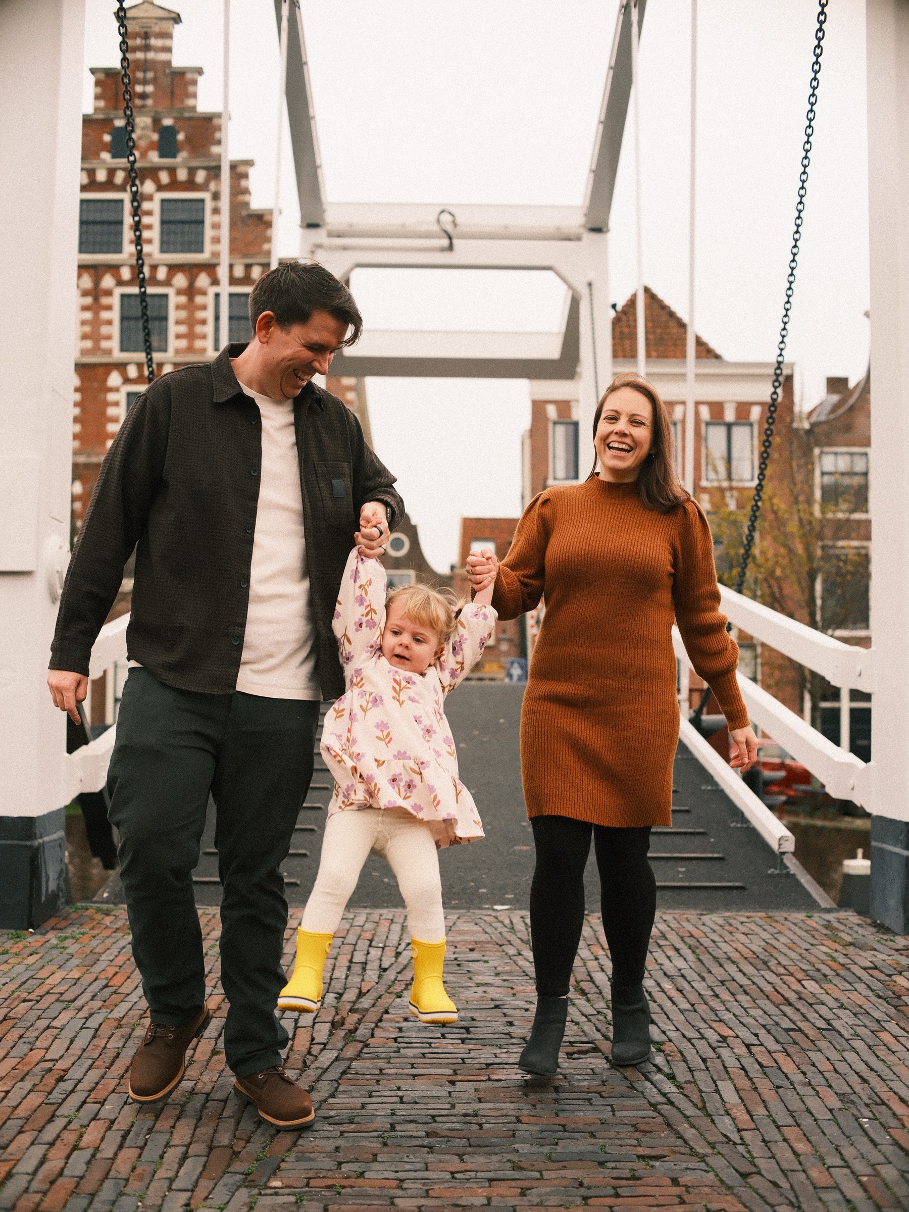 Van de witte ophaalbrug lopen de ouders met hun zwaaiende peuter tussen zich in, met een hoge trapgevel in Haarlem erachter