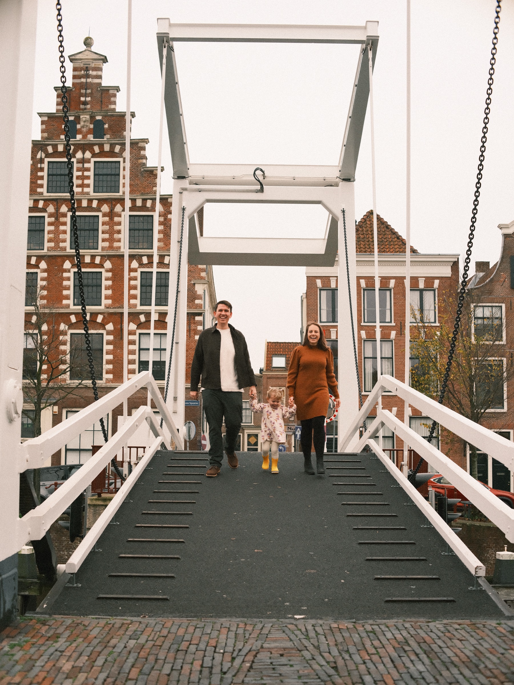 Gezin loopt over de witte ophaalbrug naar de camera toe, met de hoge witte balken en een huis met trapgevel erachter op een grijze dag