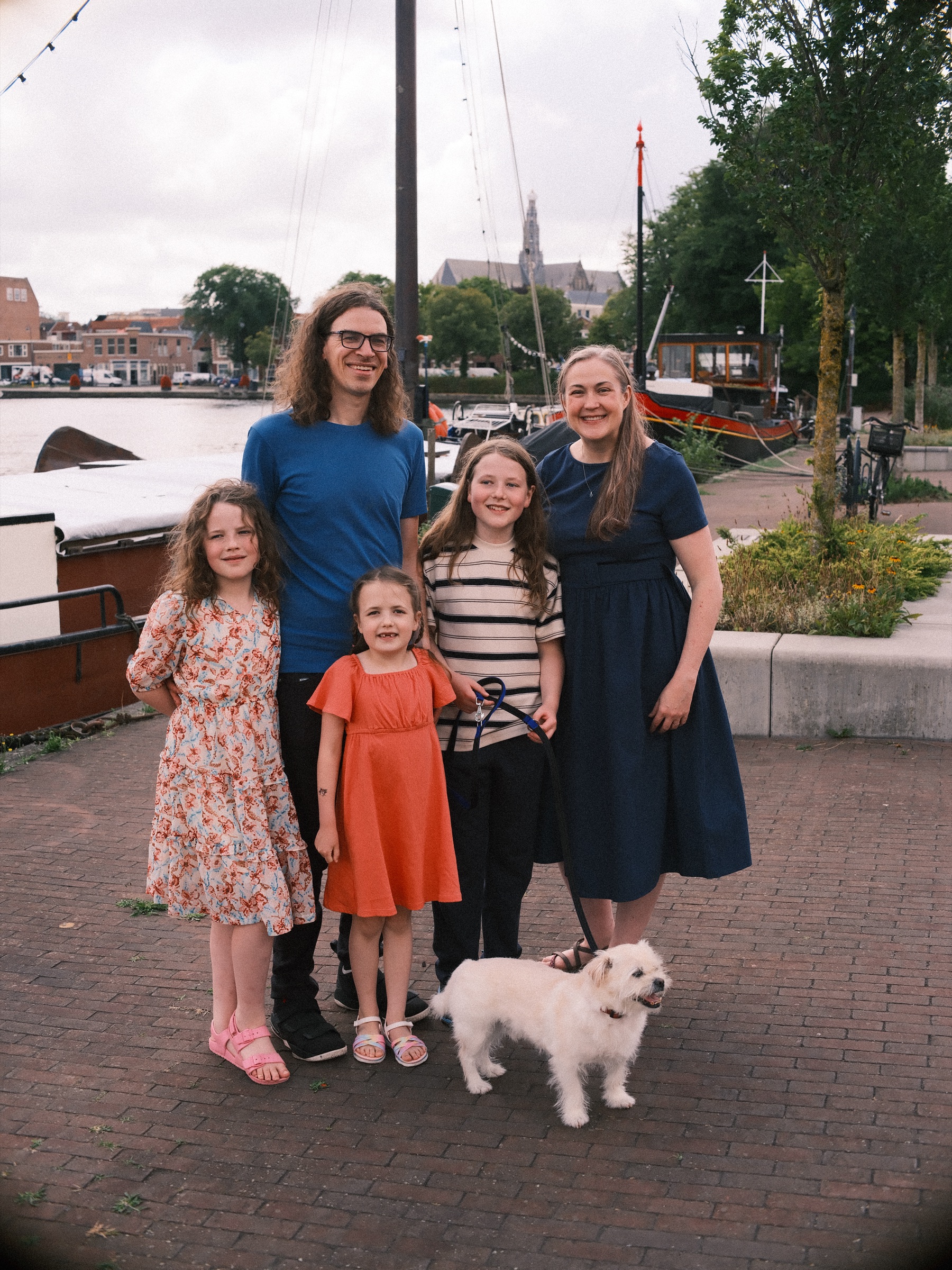 Gezin van vijf met hun kleine witte hond op een kade in Haarlem, met masten van zeilboten en een kerktoren in de verte