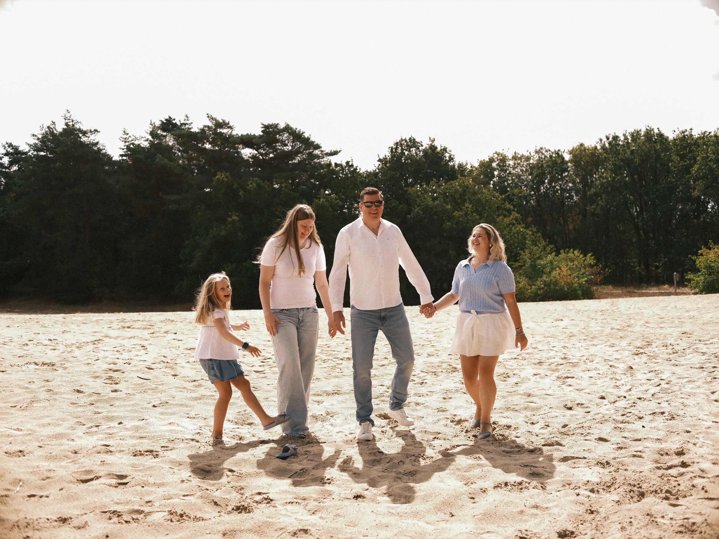 Family of four holding hands on the sand in the dunes, the youngest kicking up her leg as the three others laugh in bright sun
