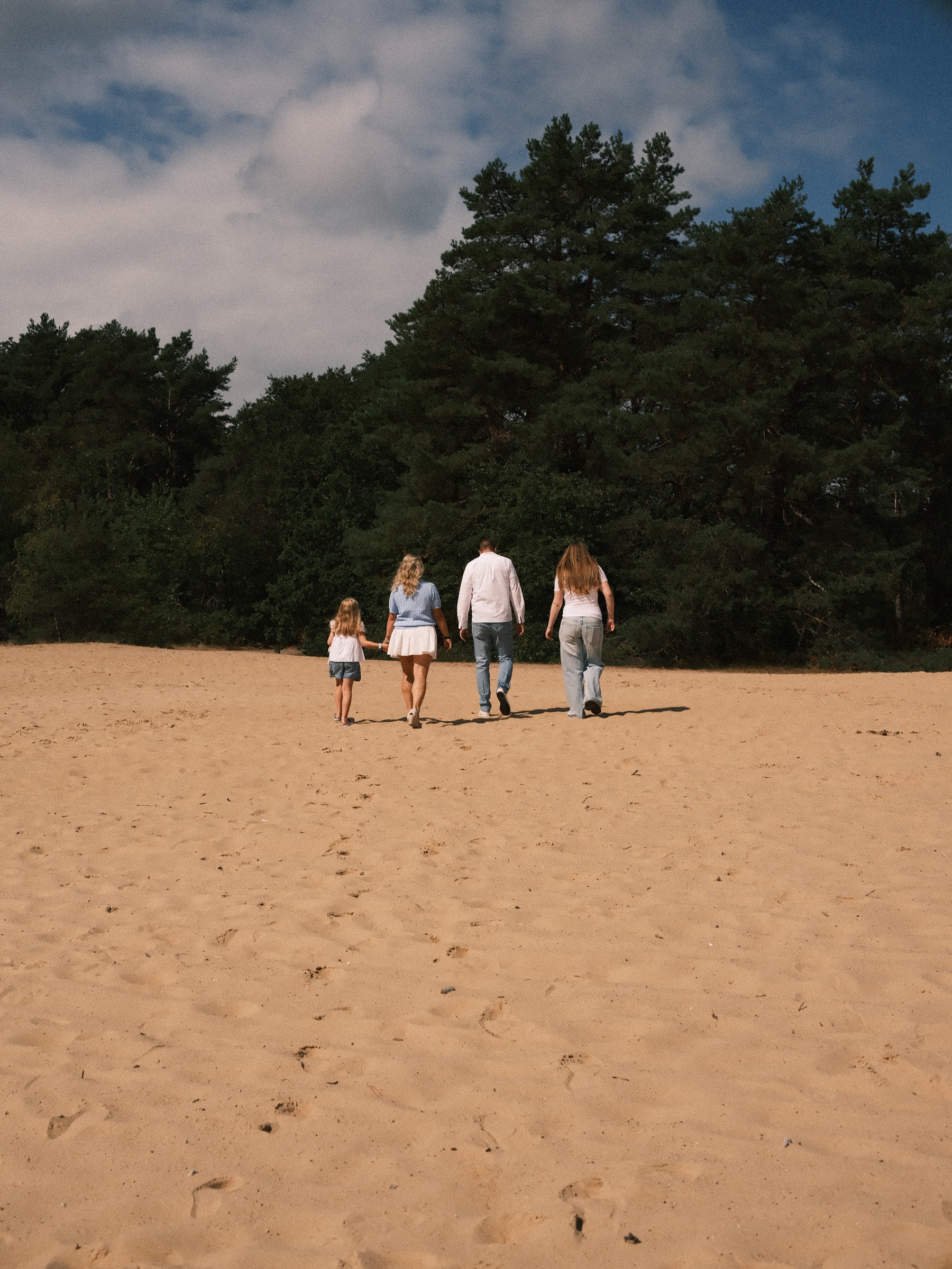Family of four walking away across the open sand toward a dark line of pines, under a blue sky with big white clouds