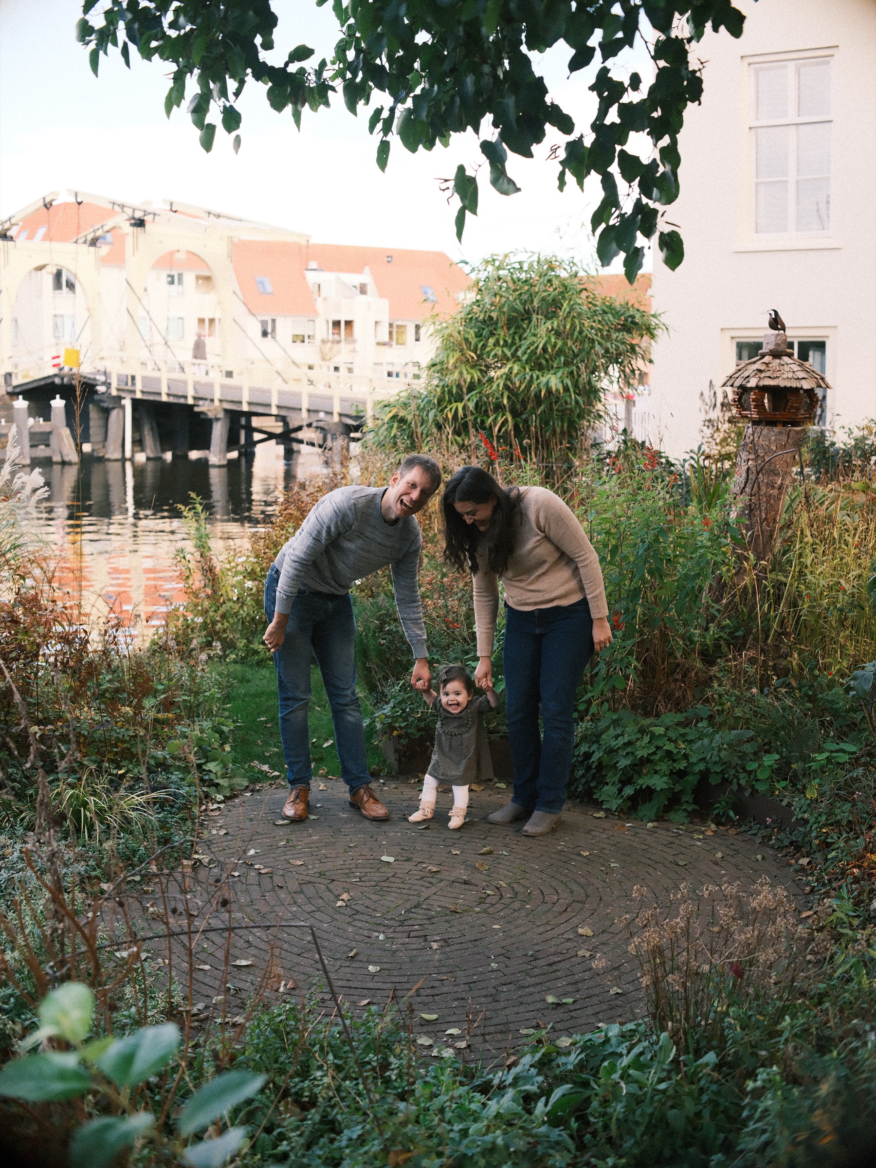 Parents helping their baby girl stand on a round brick path in a small garden by the water, the long wooden Rembrandtbrug behind them