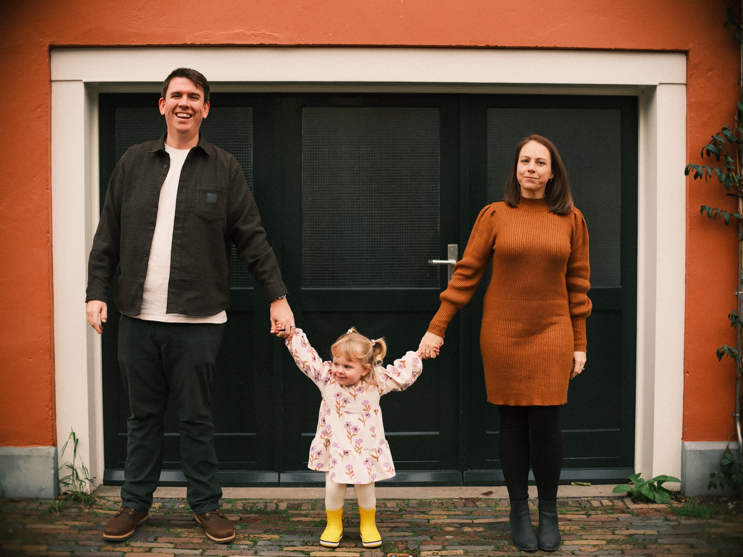 Gezin houdt elkaars handen vast voor de zwarte dubbele deuren van een oranjerood huis in Haarlem, met de peuter in het midden