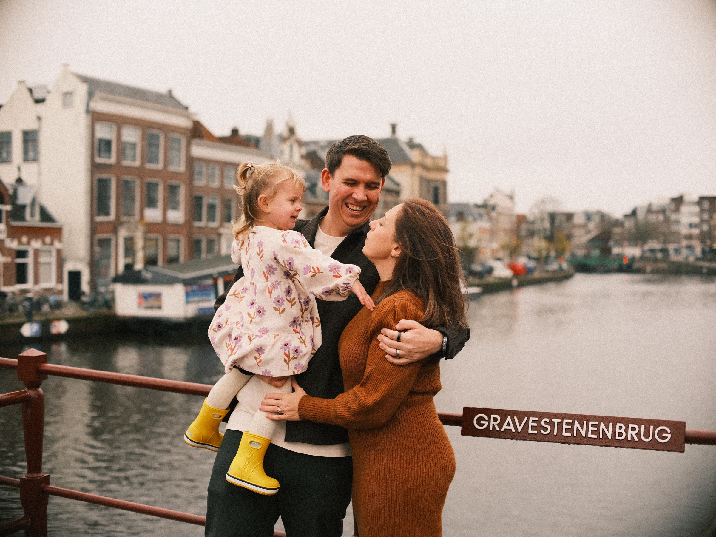 Ouders en hun peuter op gele regenlaarzen knuffelen bij de reling van de Gravestenenbrug in Haarlem, met het Spaarne erachter