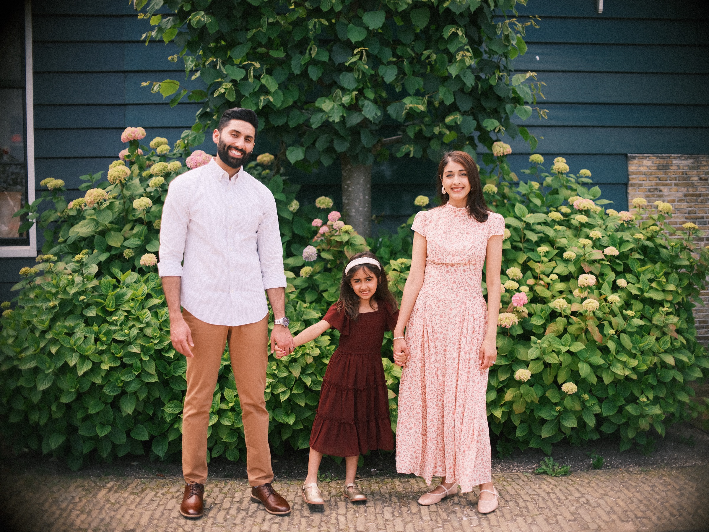 Family standing hand in hand before a wall of hydrangeas and a dark wooden house at the Zaanse Schans, the girl between her parents