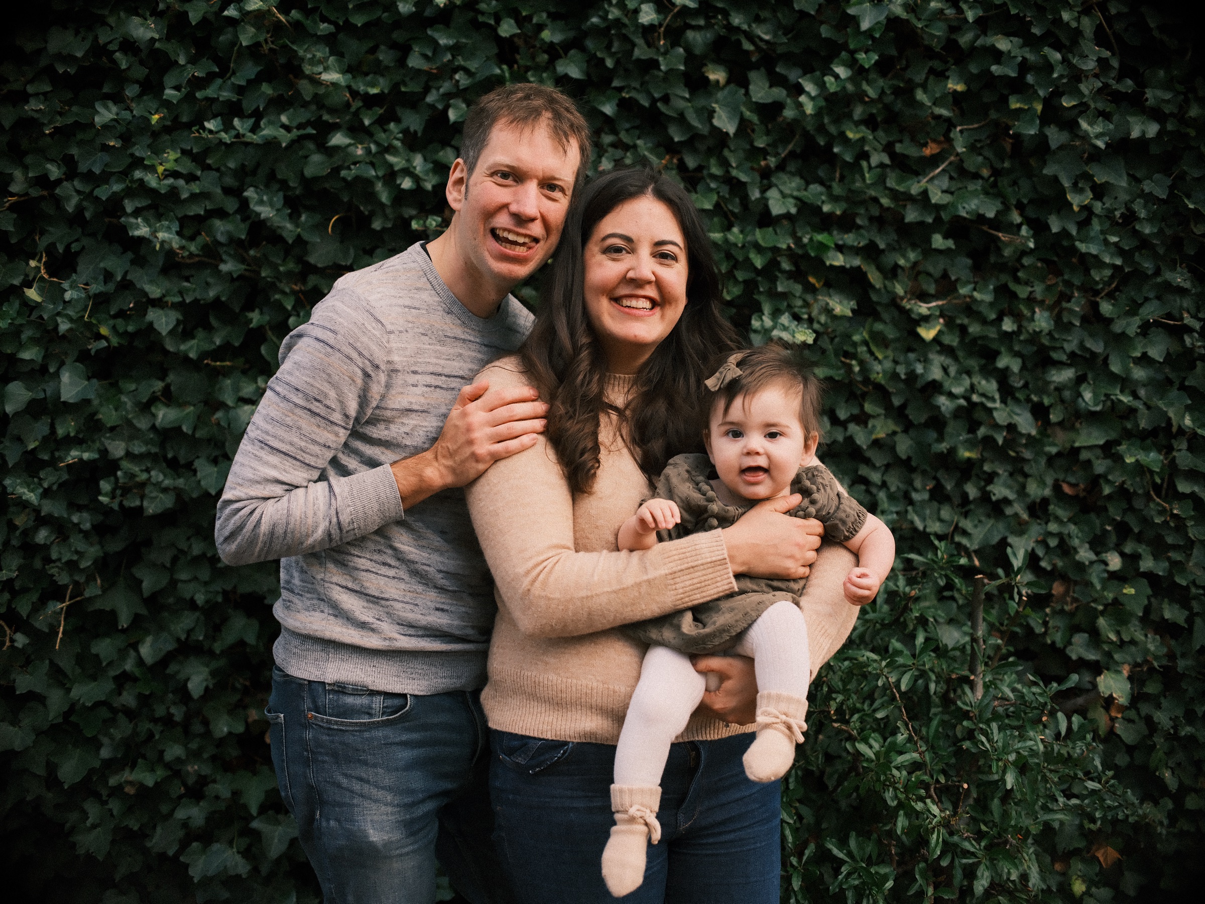 Family of three smiling in front of a dense wall of dark green ivy, the baby on her mother's hip and the father hugging them both