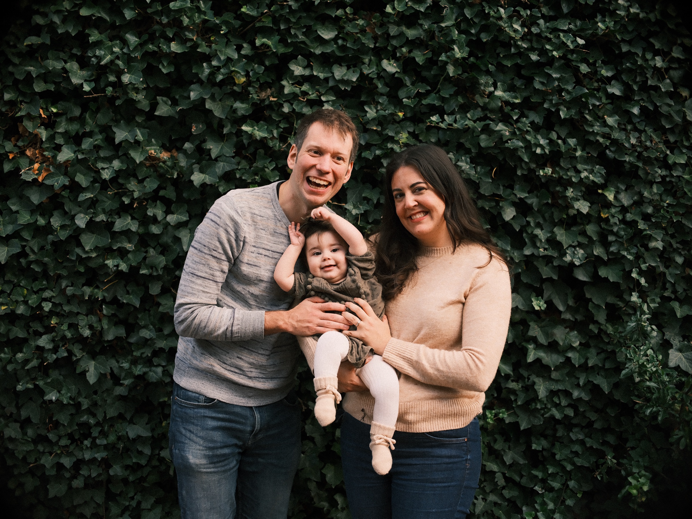 Laughing family of three before a wall of dark green ivy, the father holding up the baby girl while the mother smiles at his side