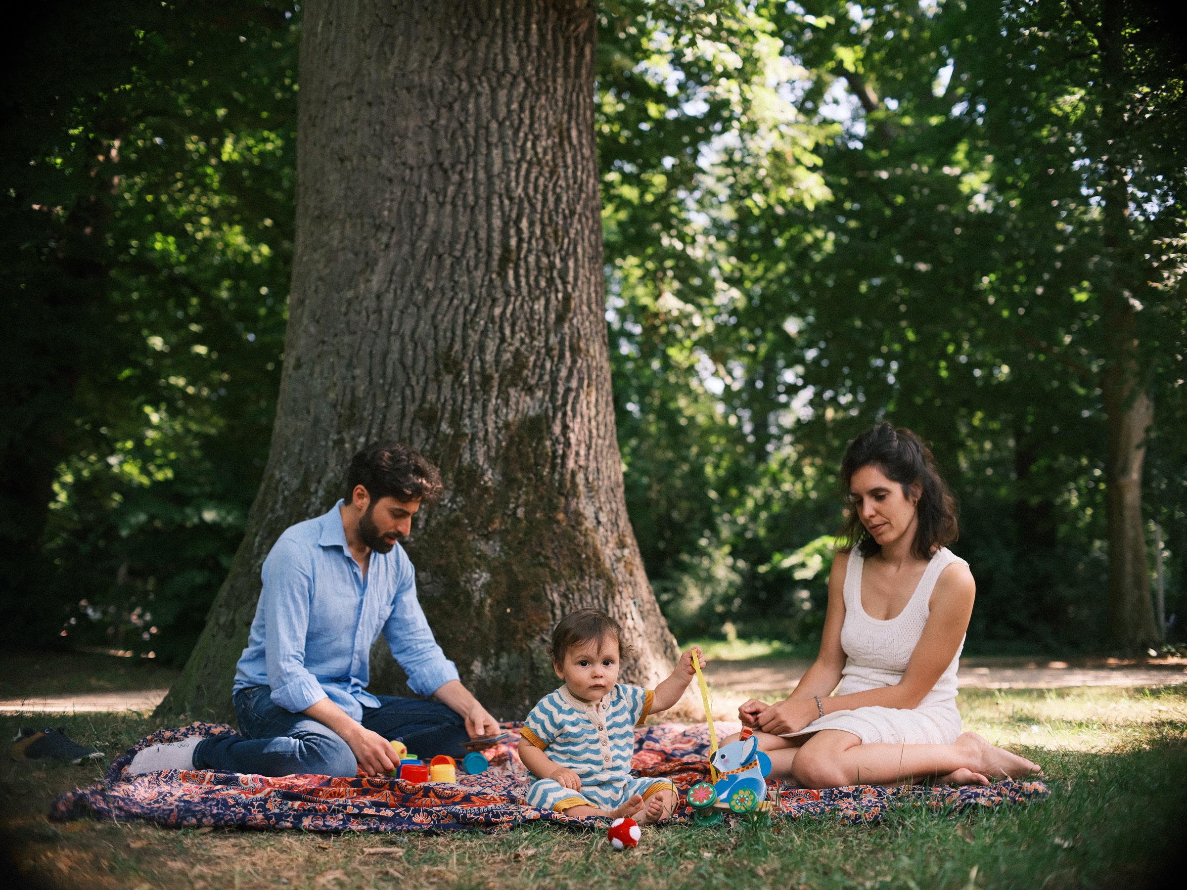 Gezin op een kleed met paisleymotief onder een grote oude boom, de baby tussen zijn ouders, tijdens een picknick in de Haarlemmerhout