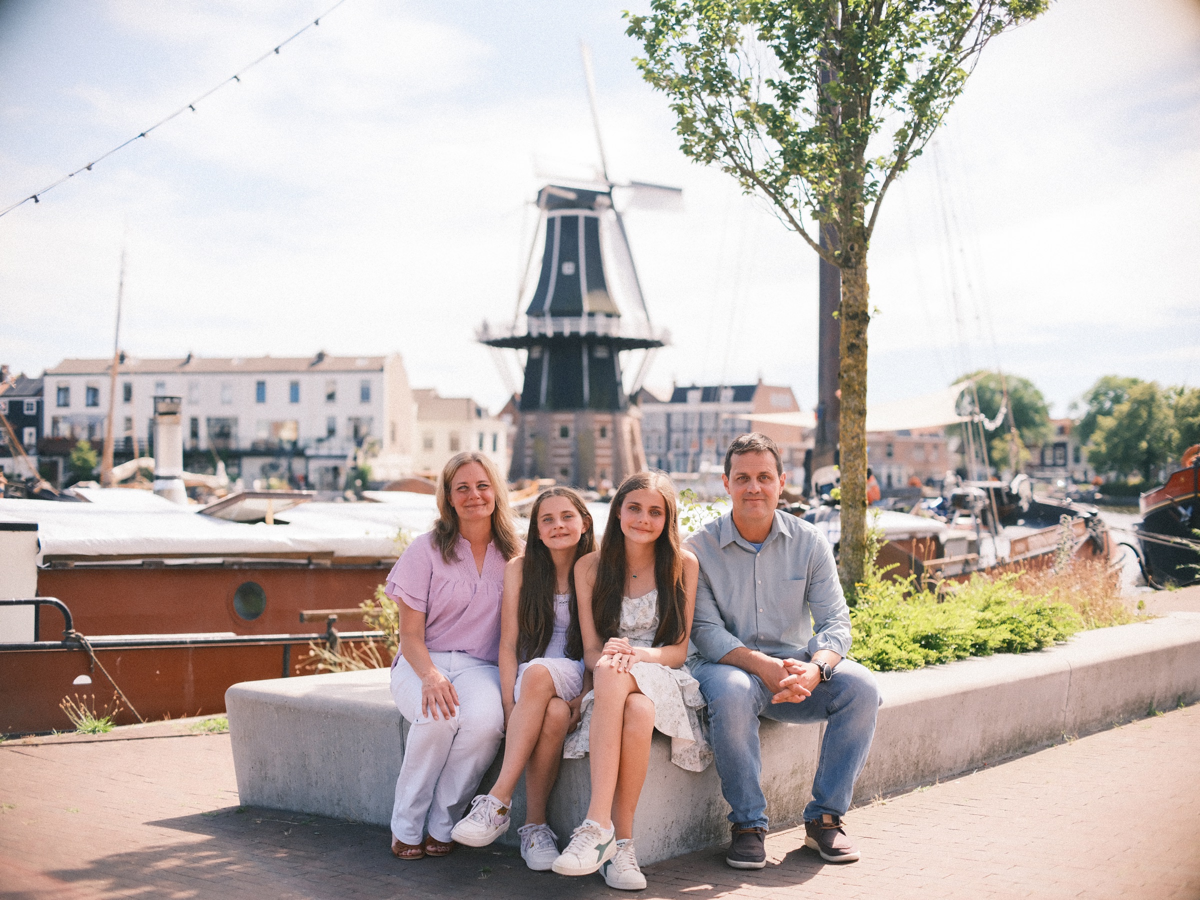 Gezin van vier naast elkaar op een zonnige betonnen plantenbak aan het Spaarne in Haarlem, met molen De Adriaan en aangemeerde boten erachter