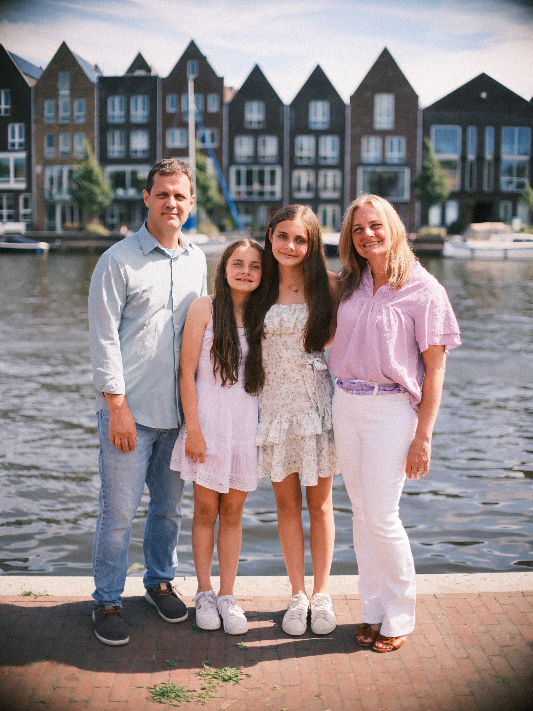 Ouders en hun twee dochters in wit en zachtblauw op de kade, met aan de overkant een rij nieuwe huizen met hoge puntgevels