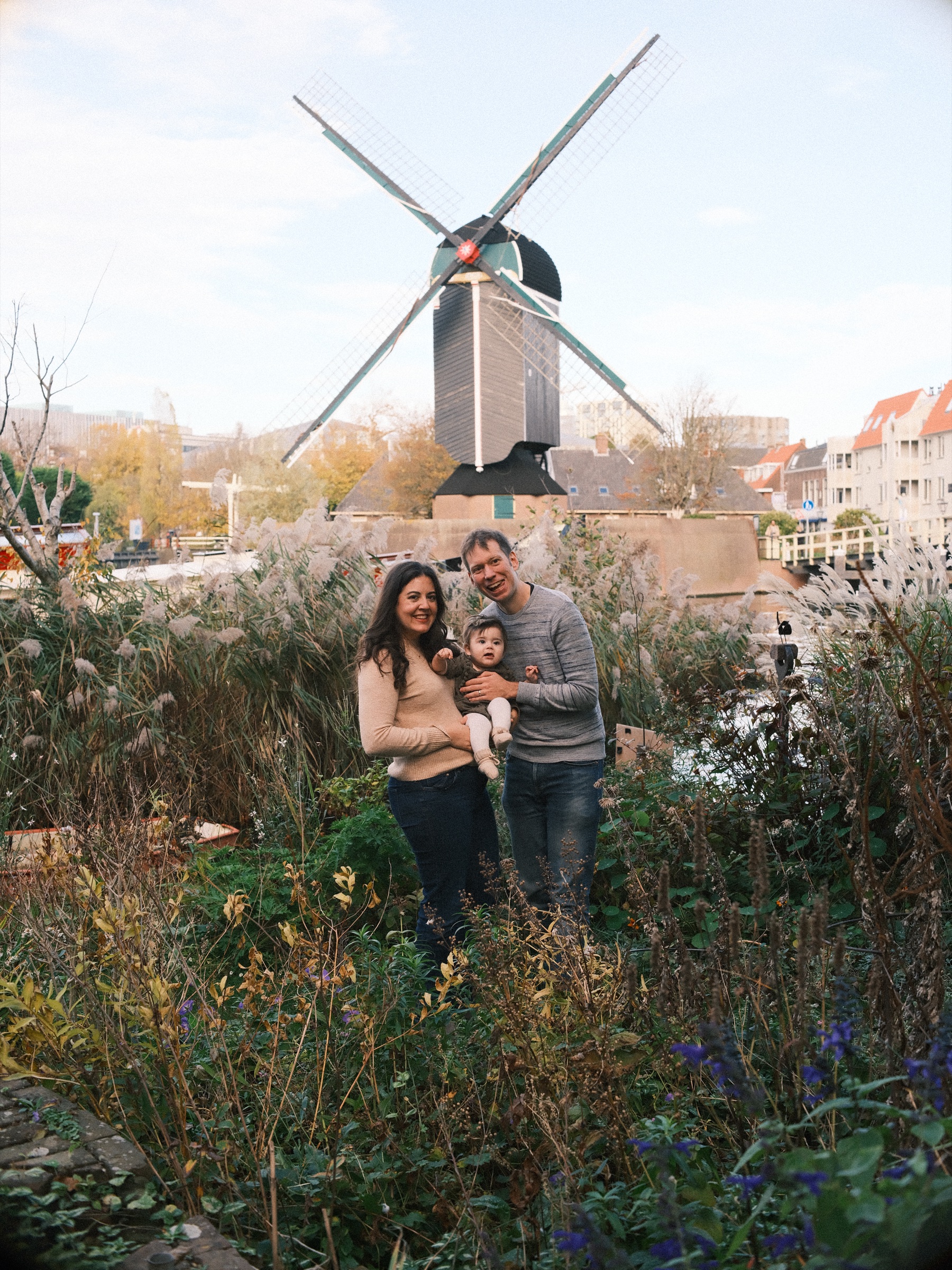 Parents holding their baby among tall grasses and reeds, Molen De Put across the water behind them and its sails against a pale autumn sky