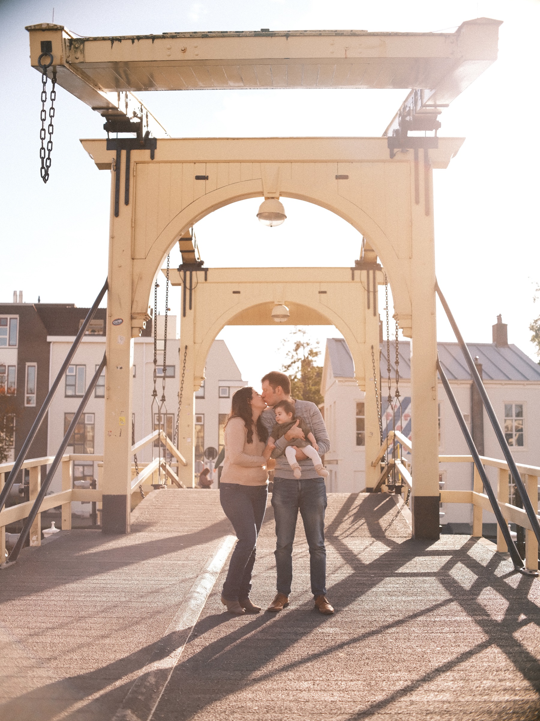 Parents kissing over their baby on the white Rembrandtbrug in Leiden, the low November sun drawing the beams of the bridge as shadows on the deck