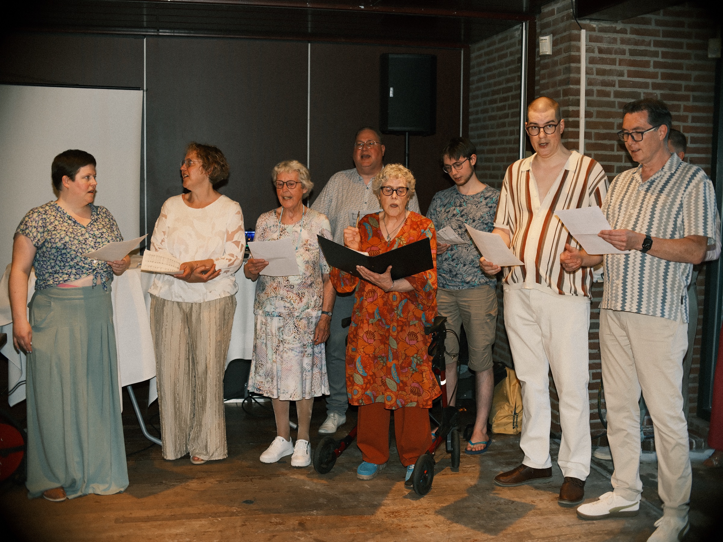 Family singing for the grooms from printed song sheets in a brick hall, an older woman in orange holding a black folder