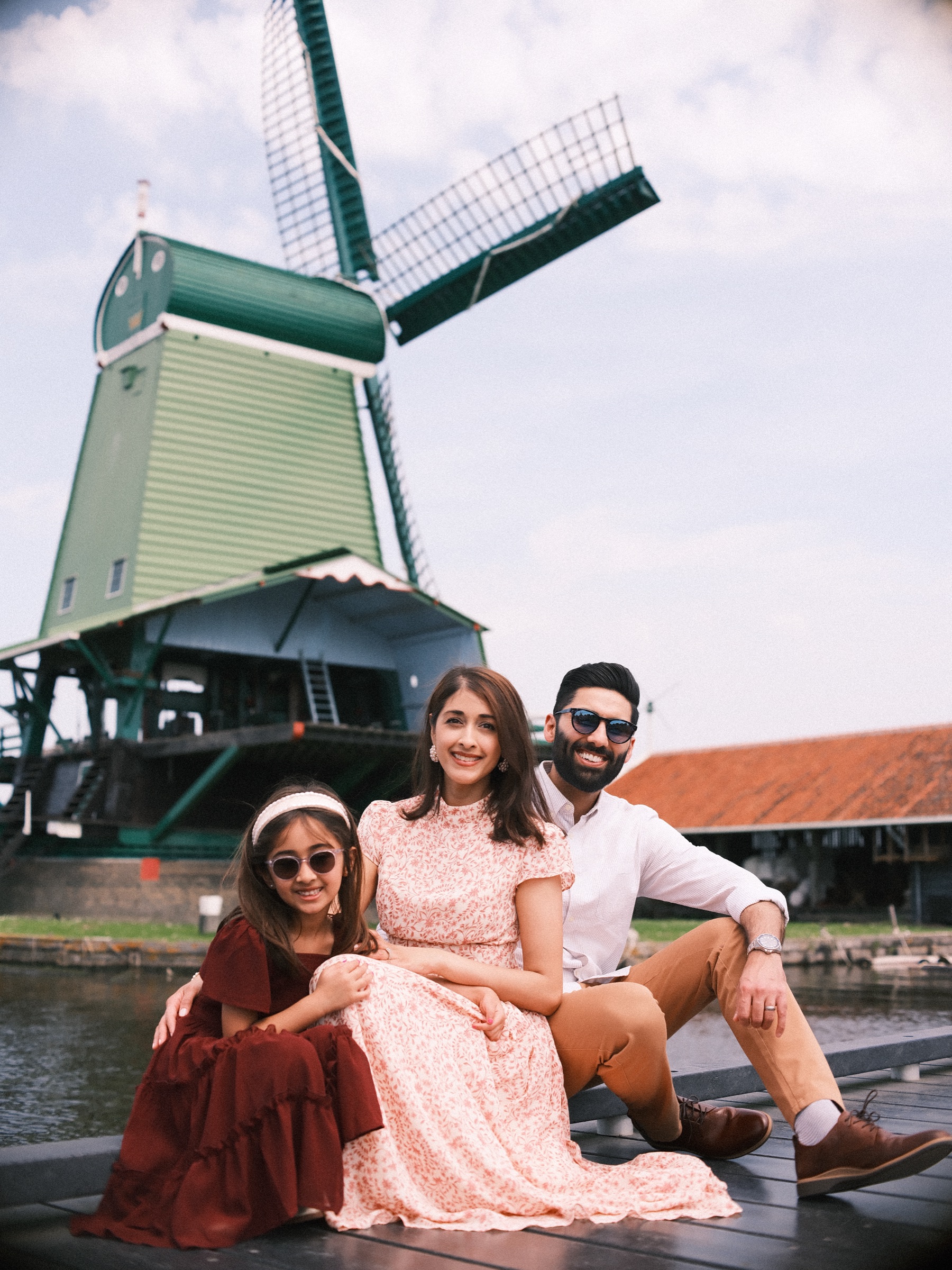 Family sitting together on the jetty under a green windmill at the Zaanse Schans, the girl in sunglasses leaning on her mother