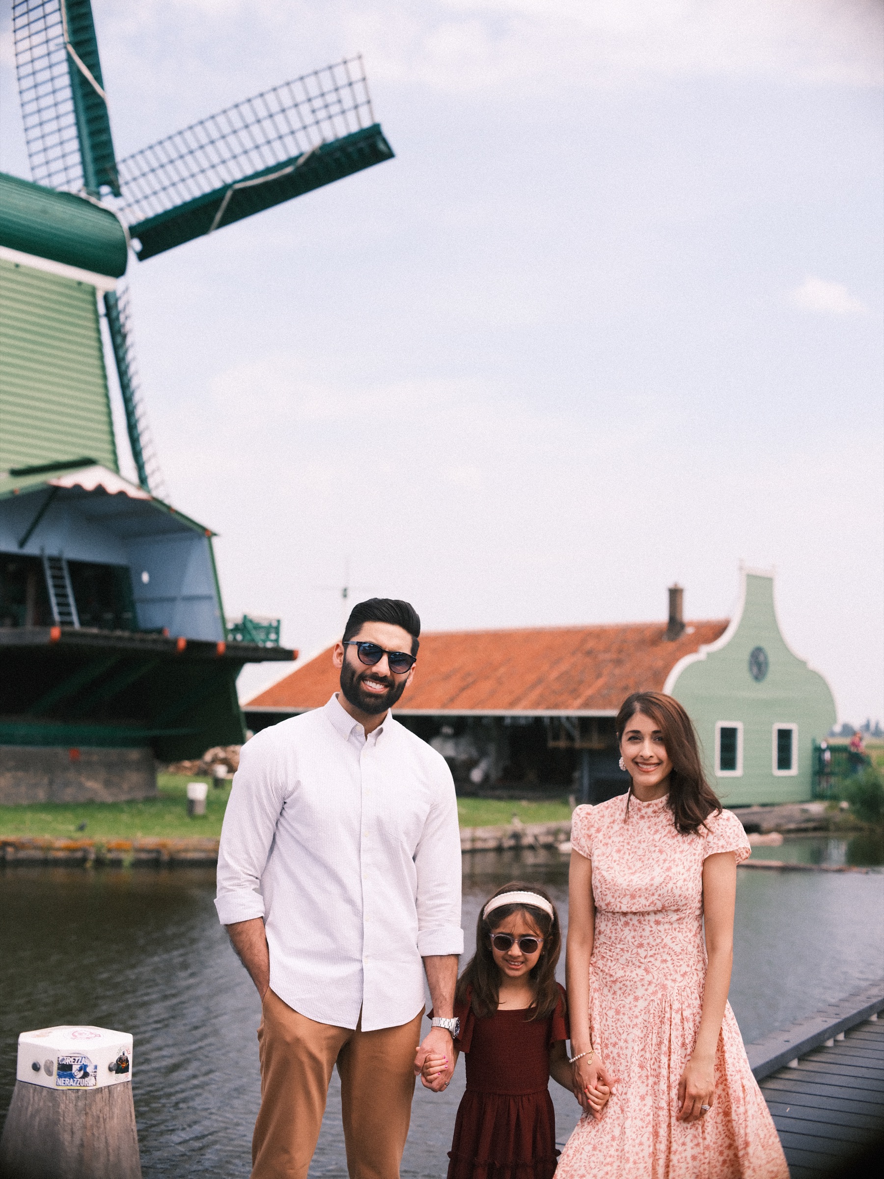 Parents and daughter standing on a wooden jetty below the big green windmill, a gabled green house and orange roofs behind them