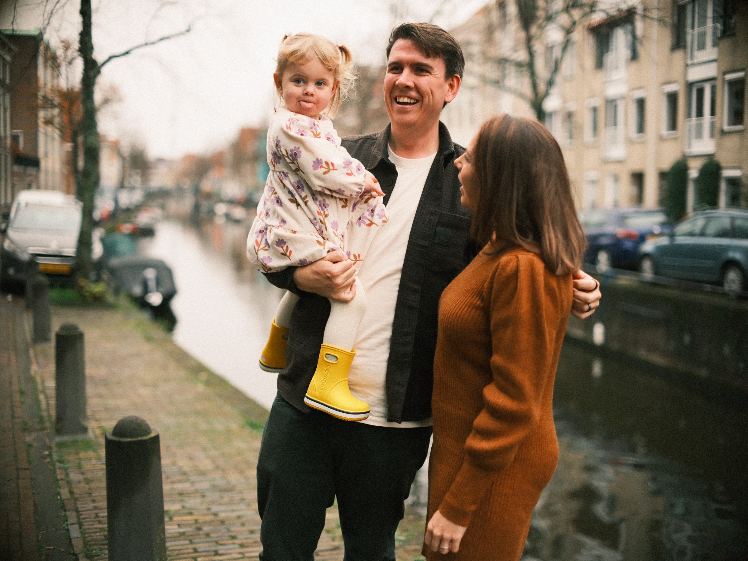 Lachende vader met zijn peuter op gele regenlaarzen op de arm, de moeder naast hen aan een stille gracht in Haarlem met kleine bootjes