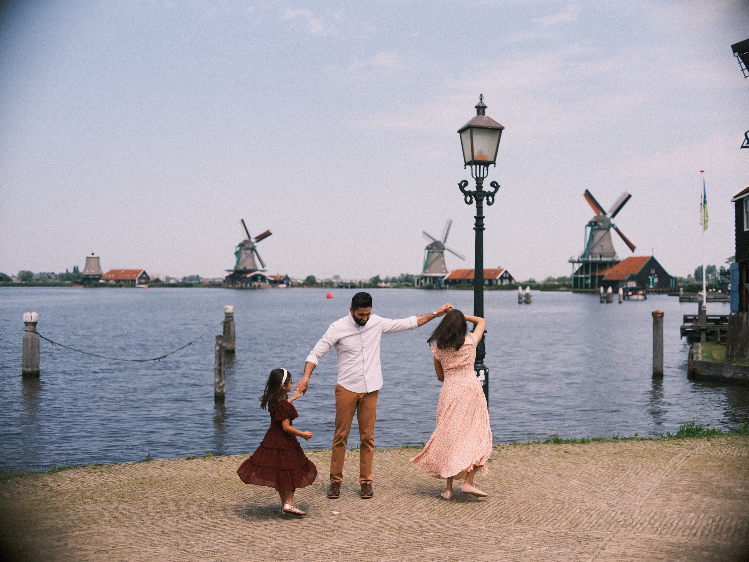 Family of three twirling hand in hand on the quay by an old lamppost, the windmills of the Zaanse Schans across the water