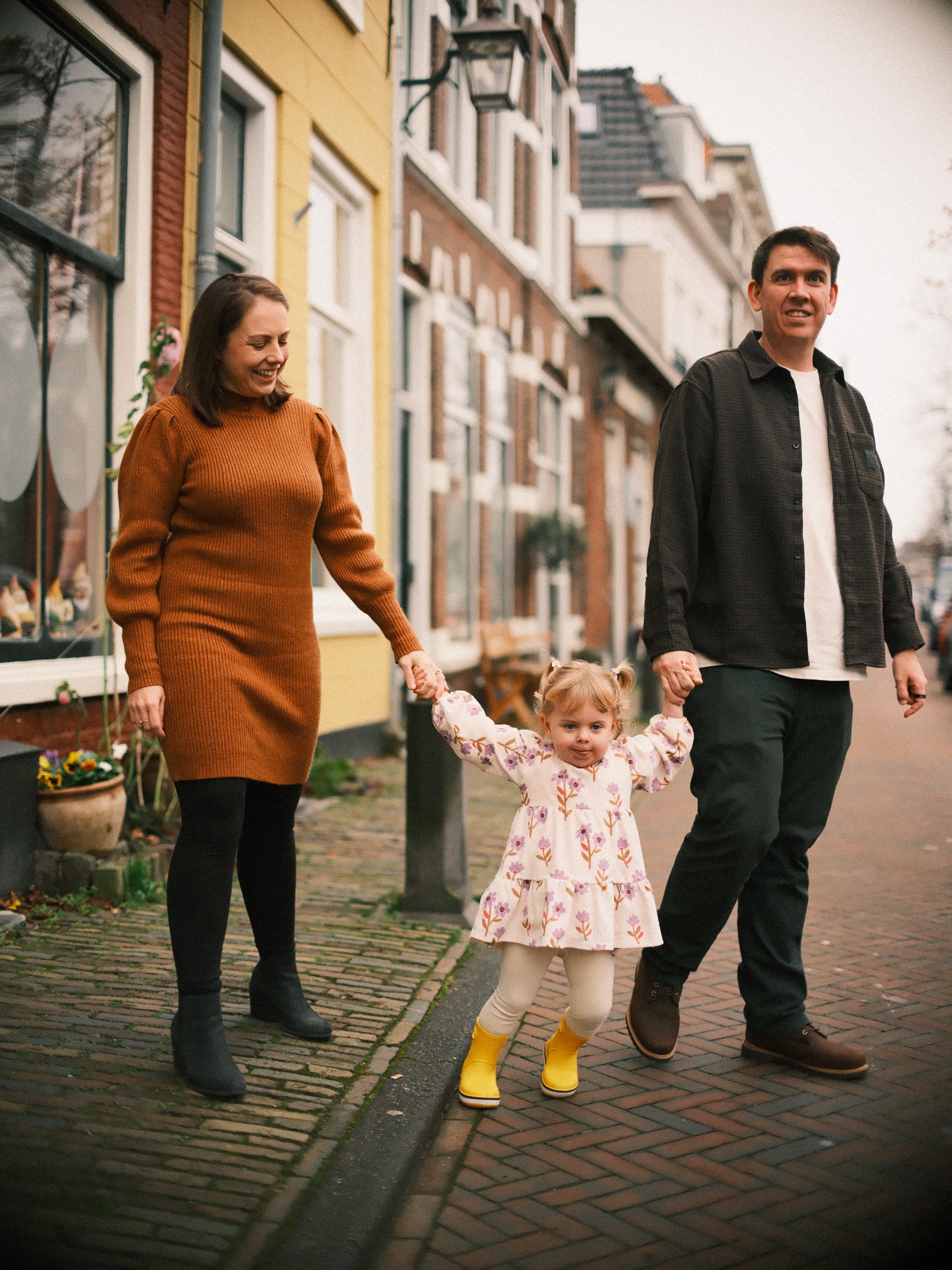 Over de klinkers lopen moeder en vader met hun dochter op gele regenlaarzen langs een geel huis en bakstenen gevels