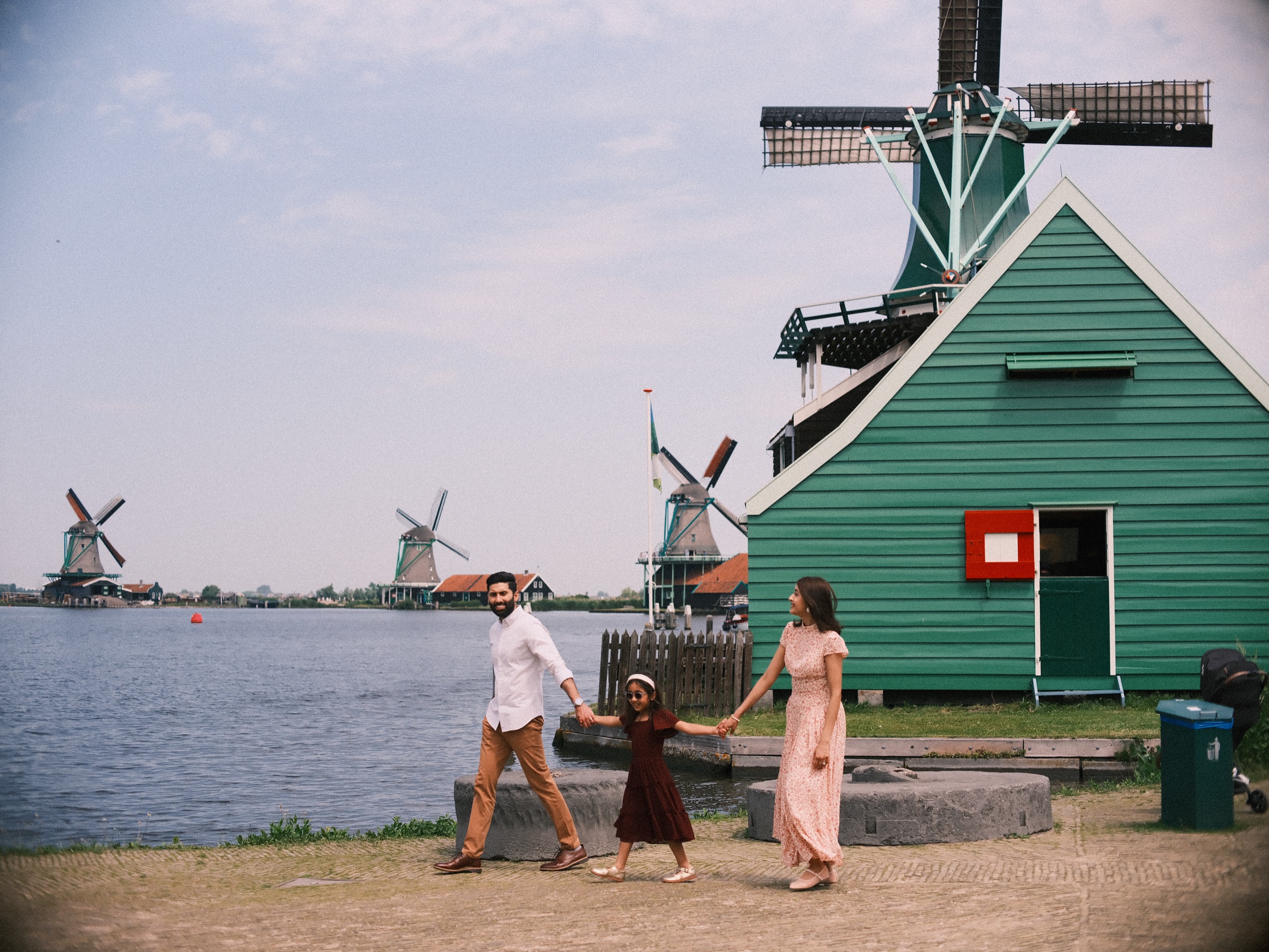 Father, mother and little daughter walking hand in hand past a green wooden mill with a red sign on its wall, the Zaan behind them