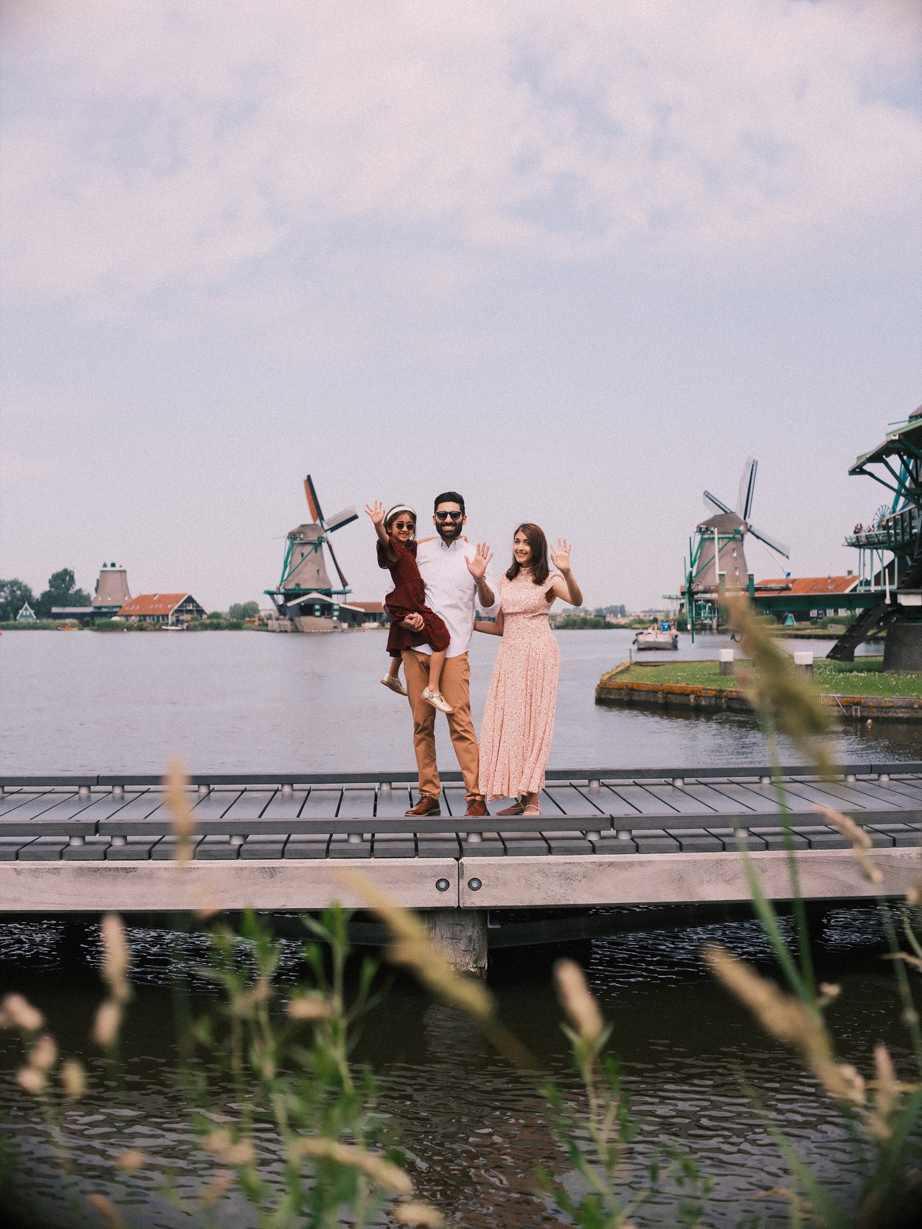 Father holding his daughter on one arm as the three of them wave from a jetty on the Zaan, windmills on the horizon and reeds in front