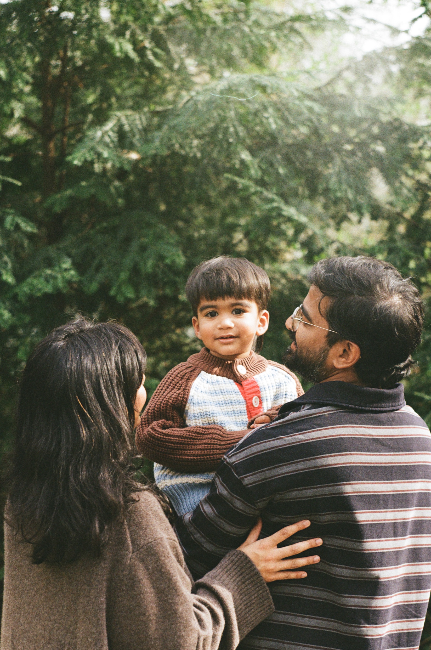 Father holding up his son while the mother looks up at the boy, dark evergreens behind them on a spring afternoon