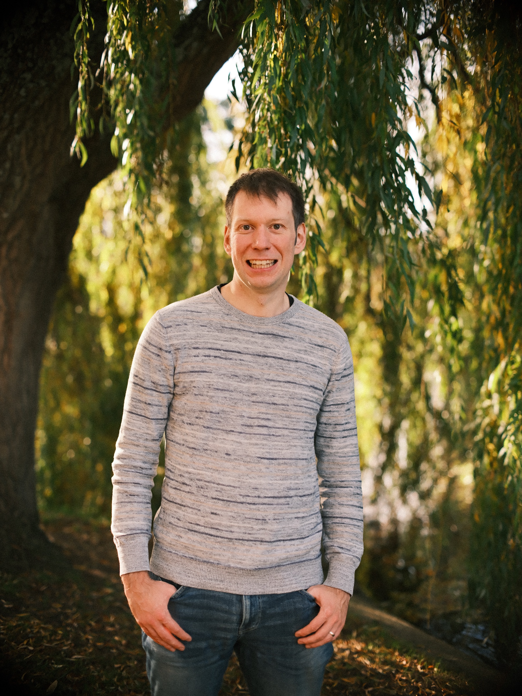 Portrait of a smiling man in a grey striped sweater with his hands in his pockets under a weeping willow in autumn in Leiden