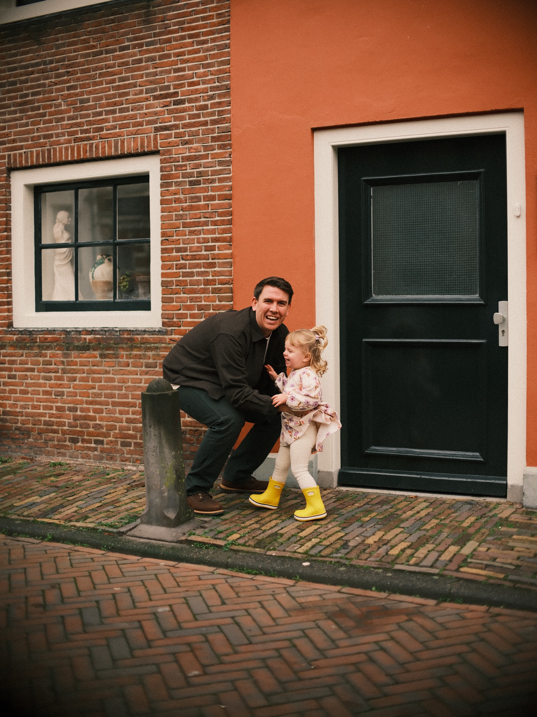 Vader zit op zijn hurken naast zijn dochtertje op gele laarzen voor een oranjerood huis met een zwarte deur en een raam met witte kozijnen