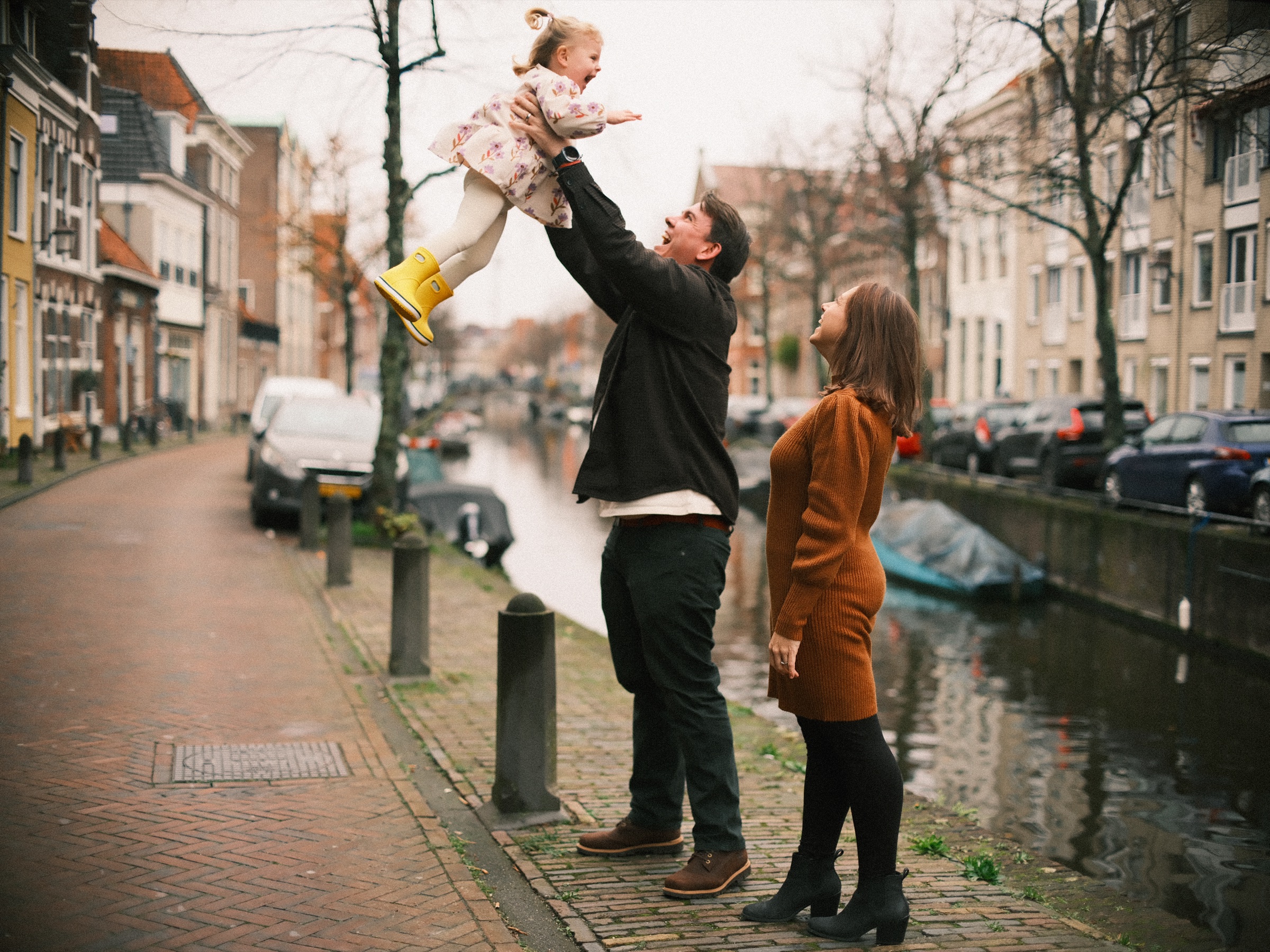 Vader gooit zijn dochter de lucht in bij een stille gracht met kale bomen en geparkeerde auto's, terwijl haar moeder toekijkt
