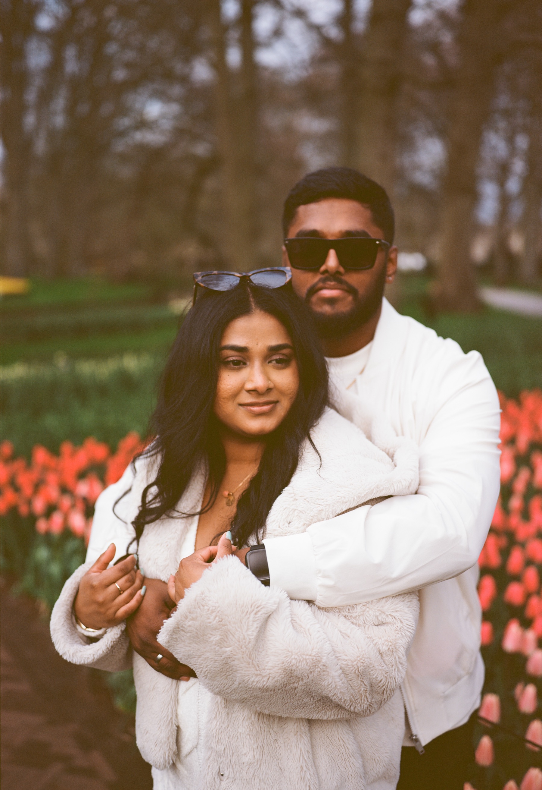 Smiling woman in a fluffy white jacket held from behind by her fiancé in dark sunglasses, red tulips around them at Keukenhof