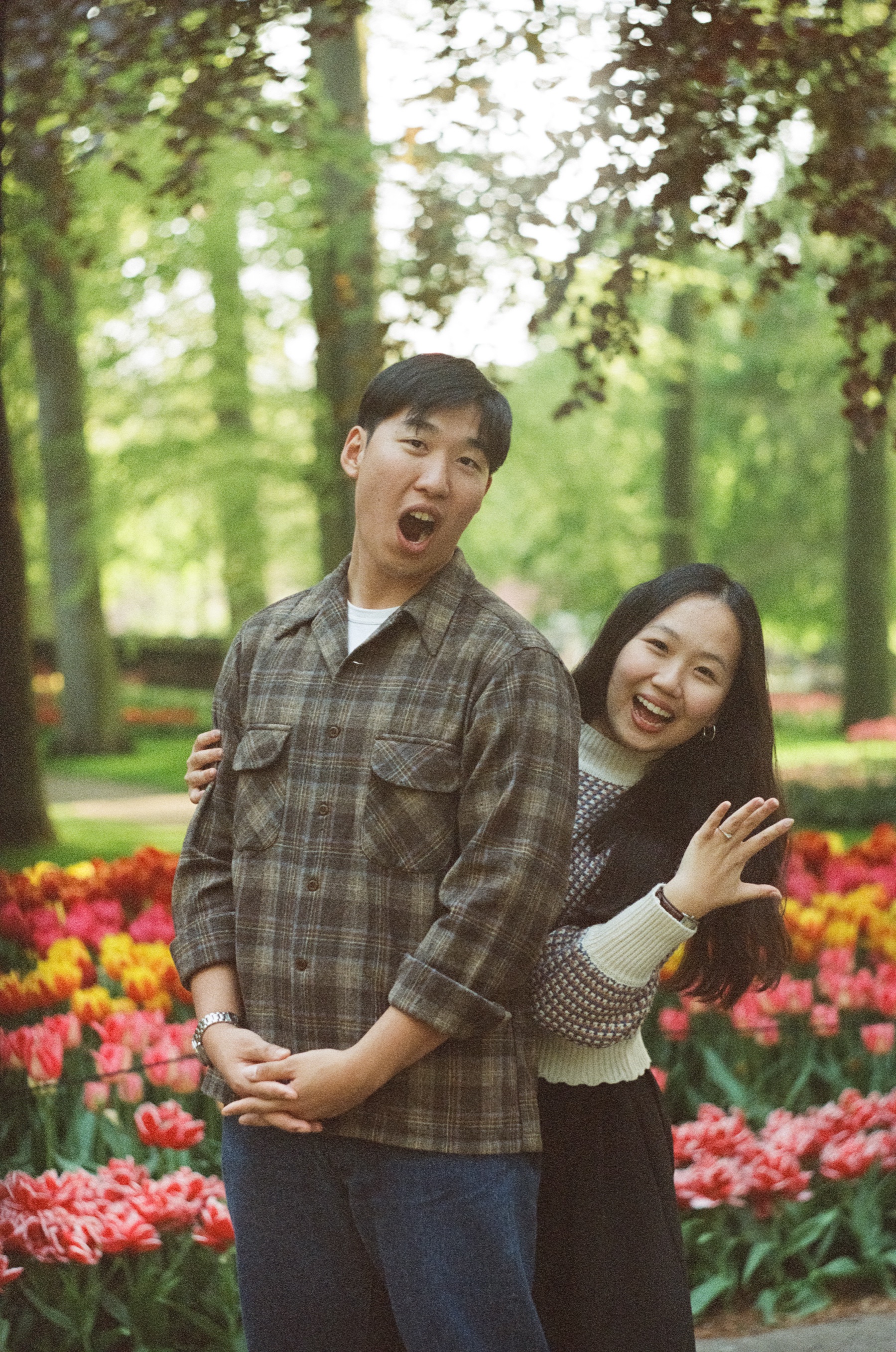 Man pulling a mock shocked face while his new fiancée laughs and shows her ring, bright tulips and trees behind them