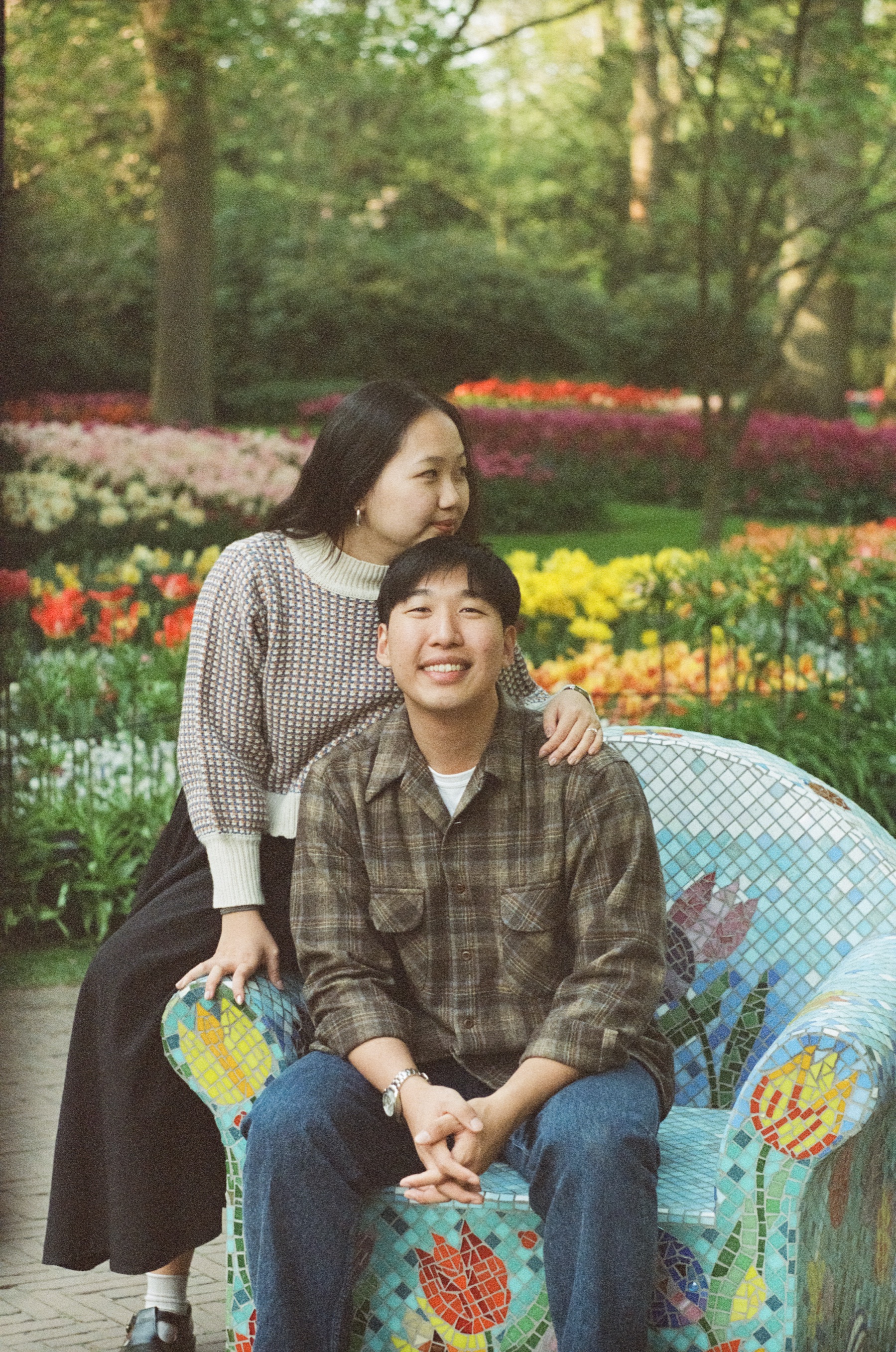 Woman leaning over her laughing fiancé from behind as he sits on a chair covered in colorful mosaic tulips