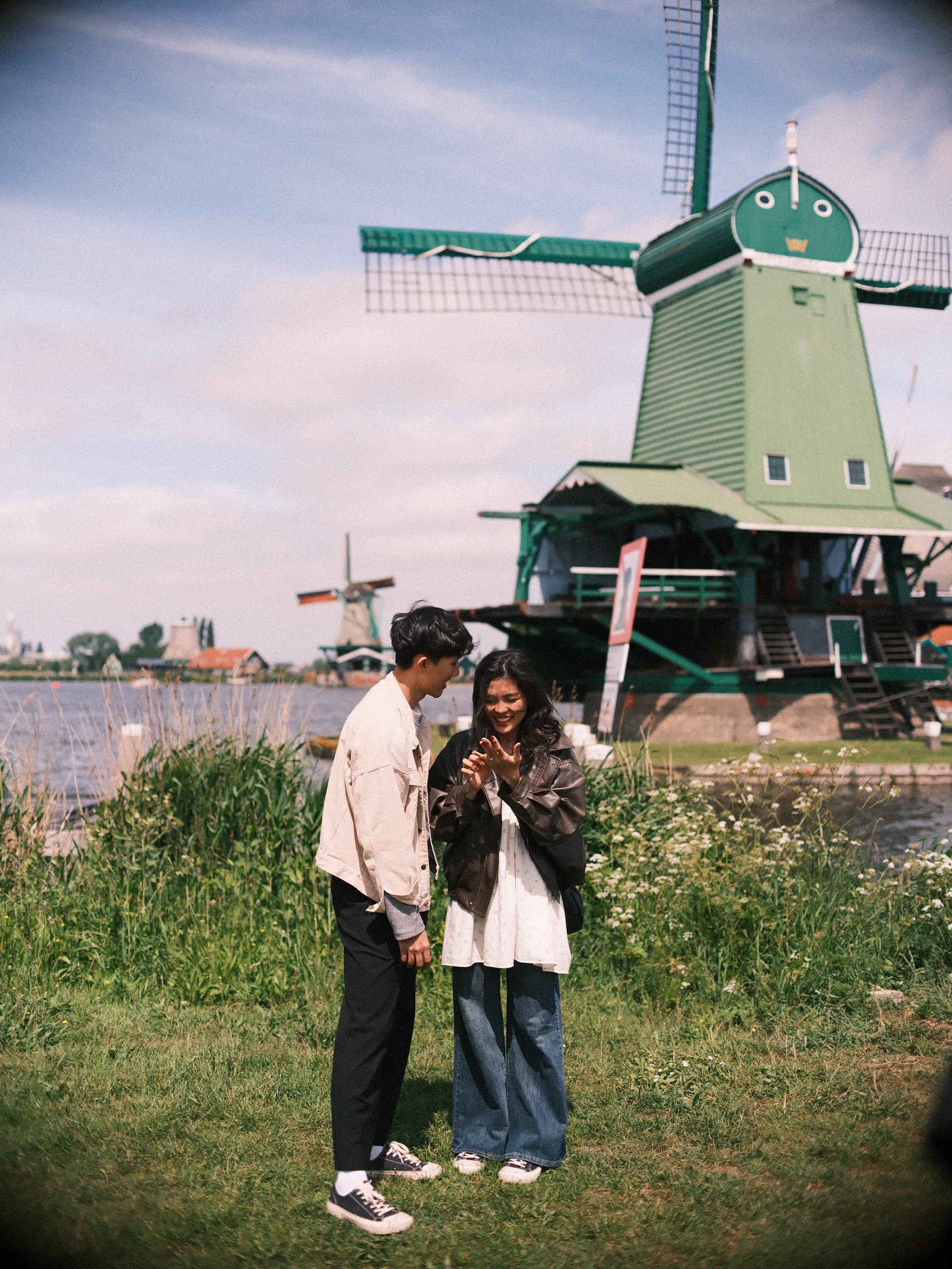 Woman in a brown leather jacket smiling at the ring on her hand while her fiancé watches, a green windmill at the Zaanse Schans behind