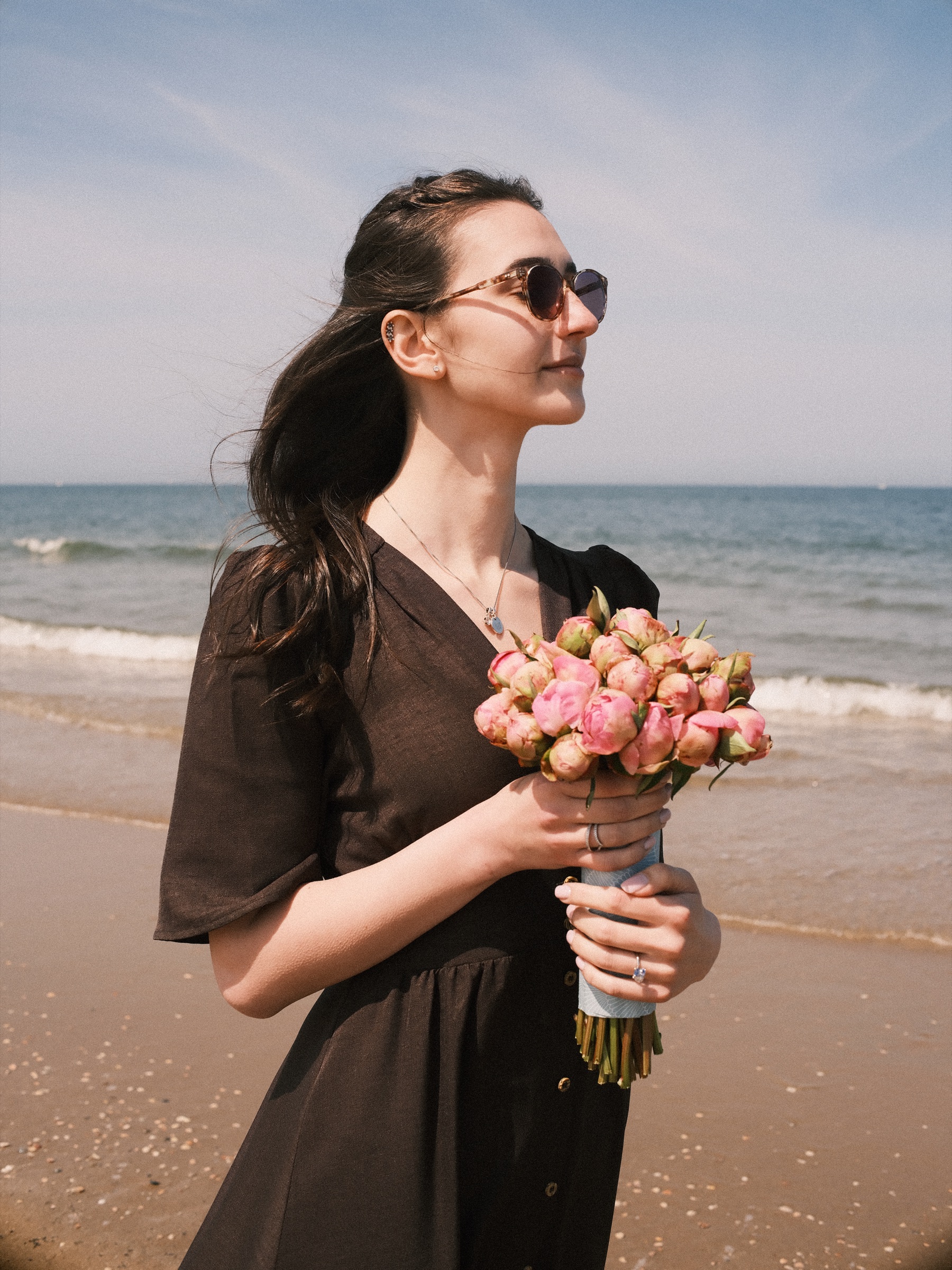 Newly engaged woman in sunglasses holding pink peonies on Scheveningen beach, her dark hair blowing in the wind