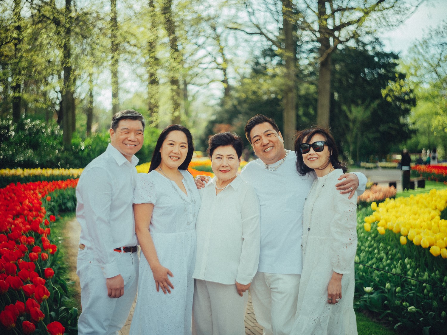 Vijf volwassenen van drie generaties in het wit staan arm in arm tussen rode en gele tulpen, met oma in het midden