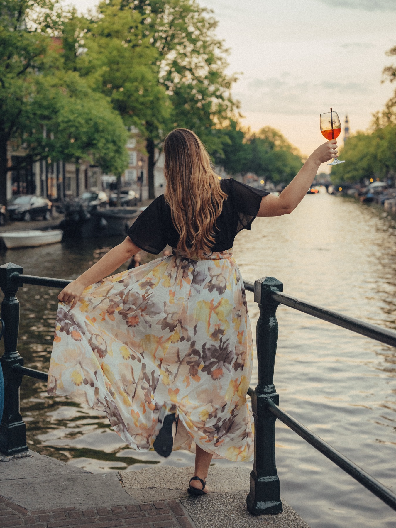 Friend in a floral maxi skirt raising her spritz toward the canal at sunset, holding her skirt out on the edge of the bridge