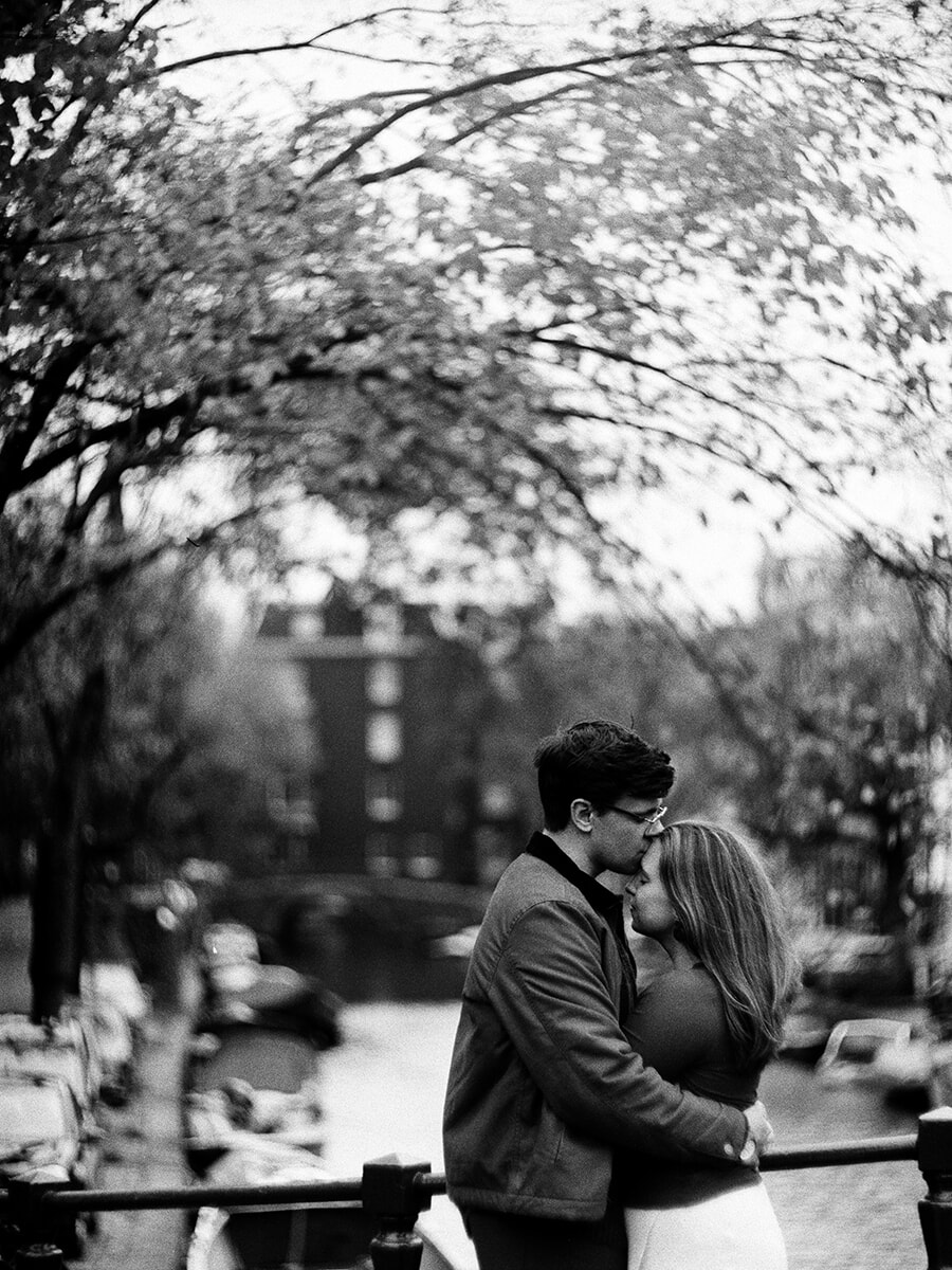 Under leafy branches, a man in glasses kisses his partner’s forehead beside a canal railing in a black-and-white portrait