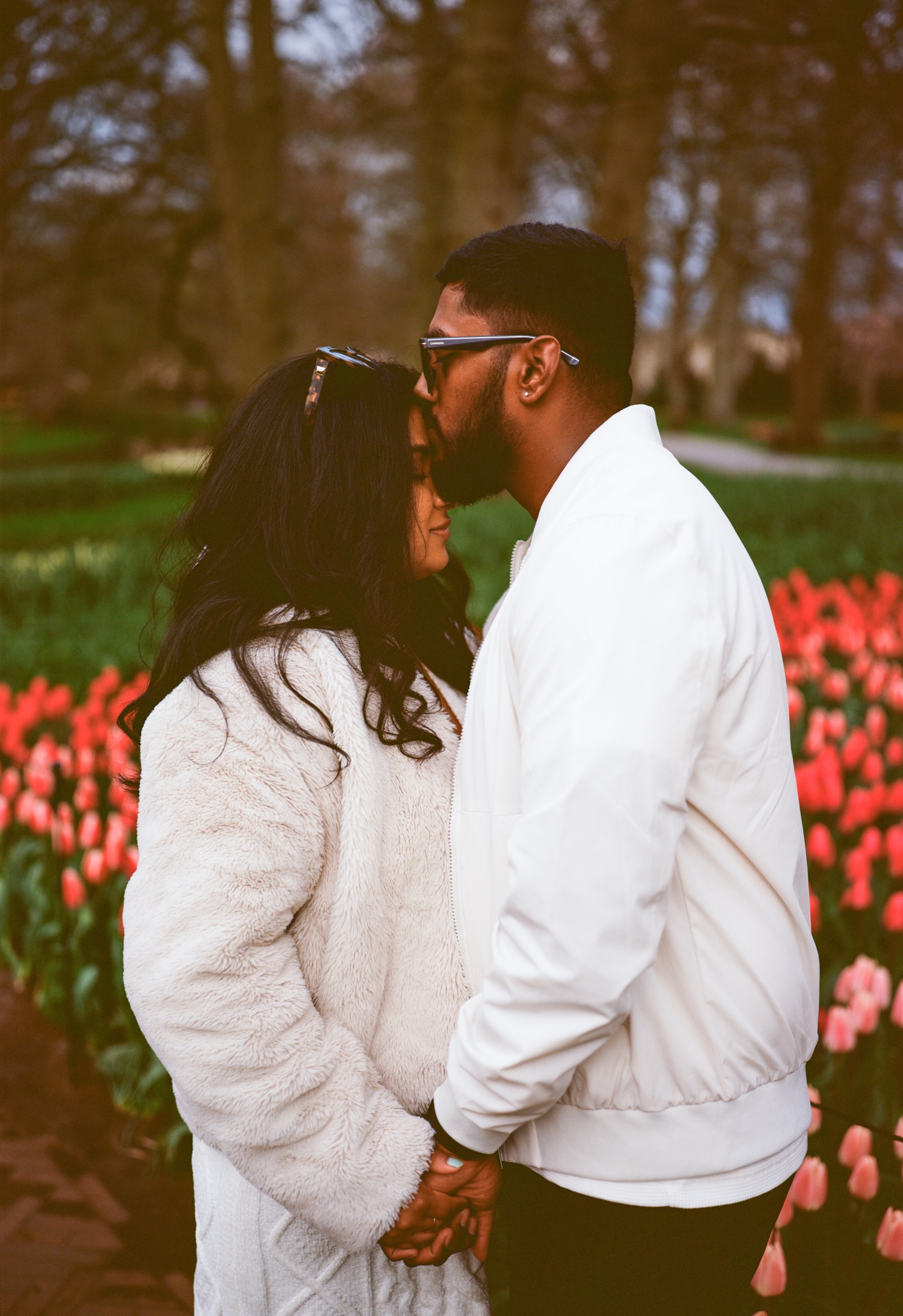 Man in sunglasses and a white jacket kissing the forehead of his new fiancée among coral-red tulips at Keukenhof