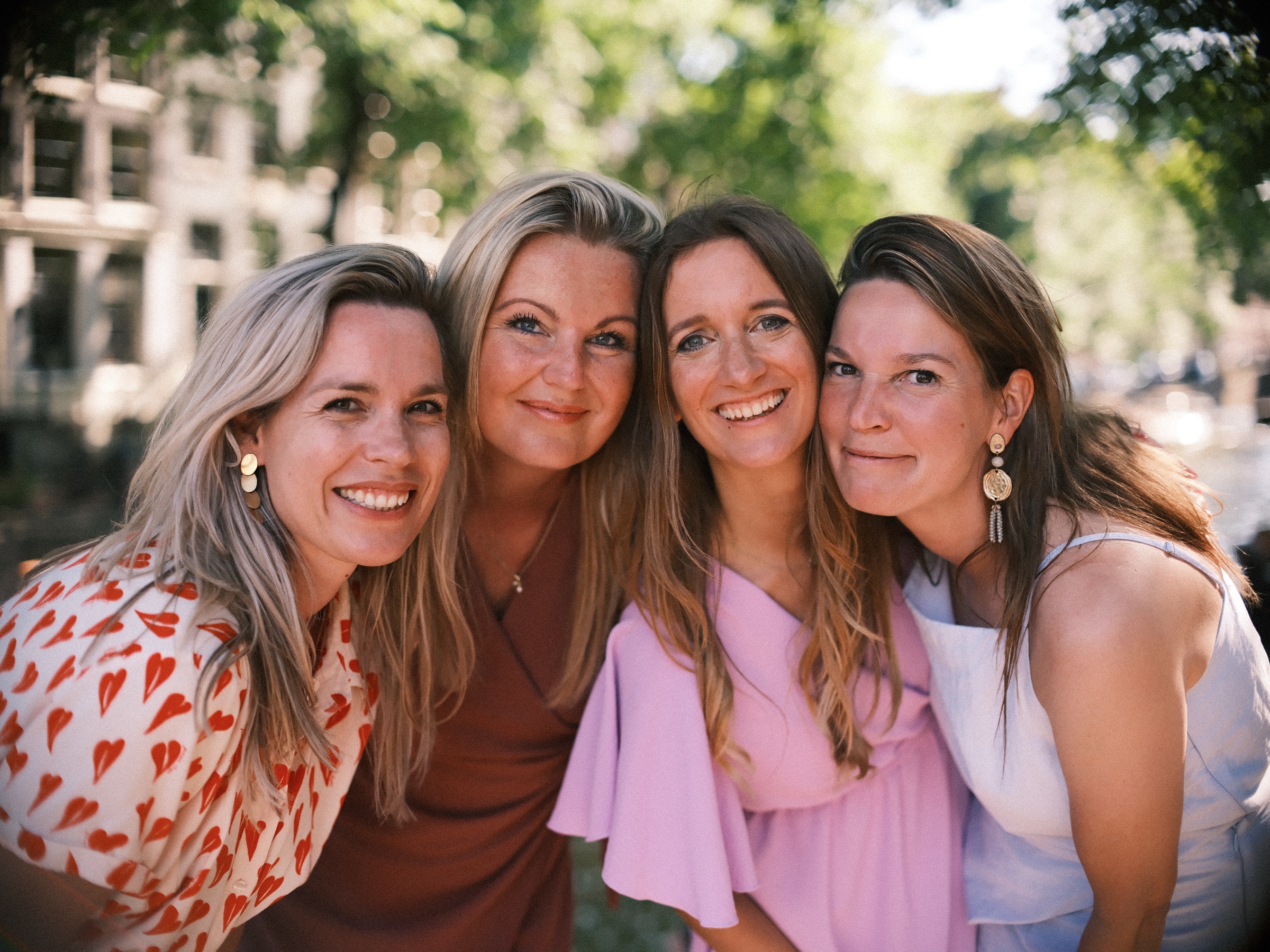 Four friends at a bachelorette pressing their heads together by a canal, all smiling, the canal houses soft behind them