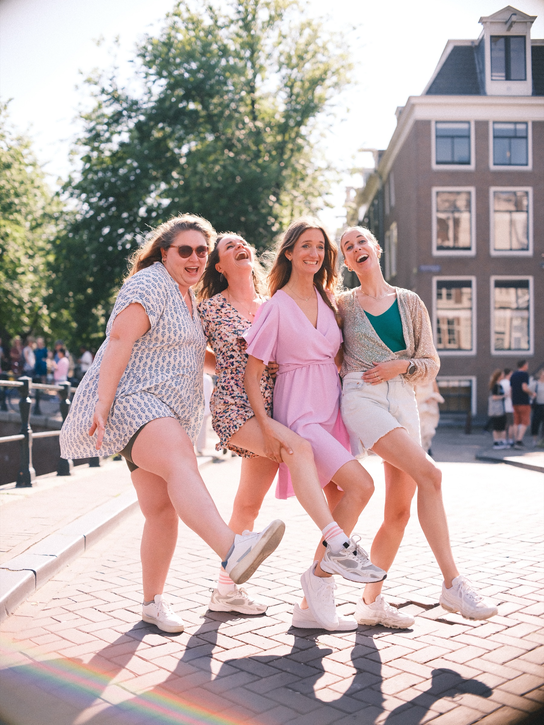 Four women laughing on a sunny canal bridge, each kicking out one leg at the same time, the bride-to-be in lilac among them