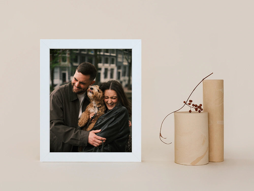White wooden frame with a laughing couple hugging their dog on an Amsterdam street, next to two wooden vases and a dried branch