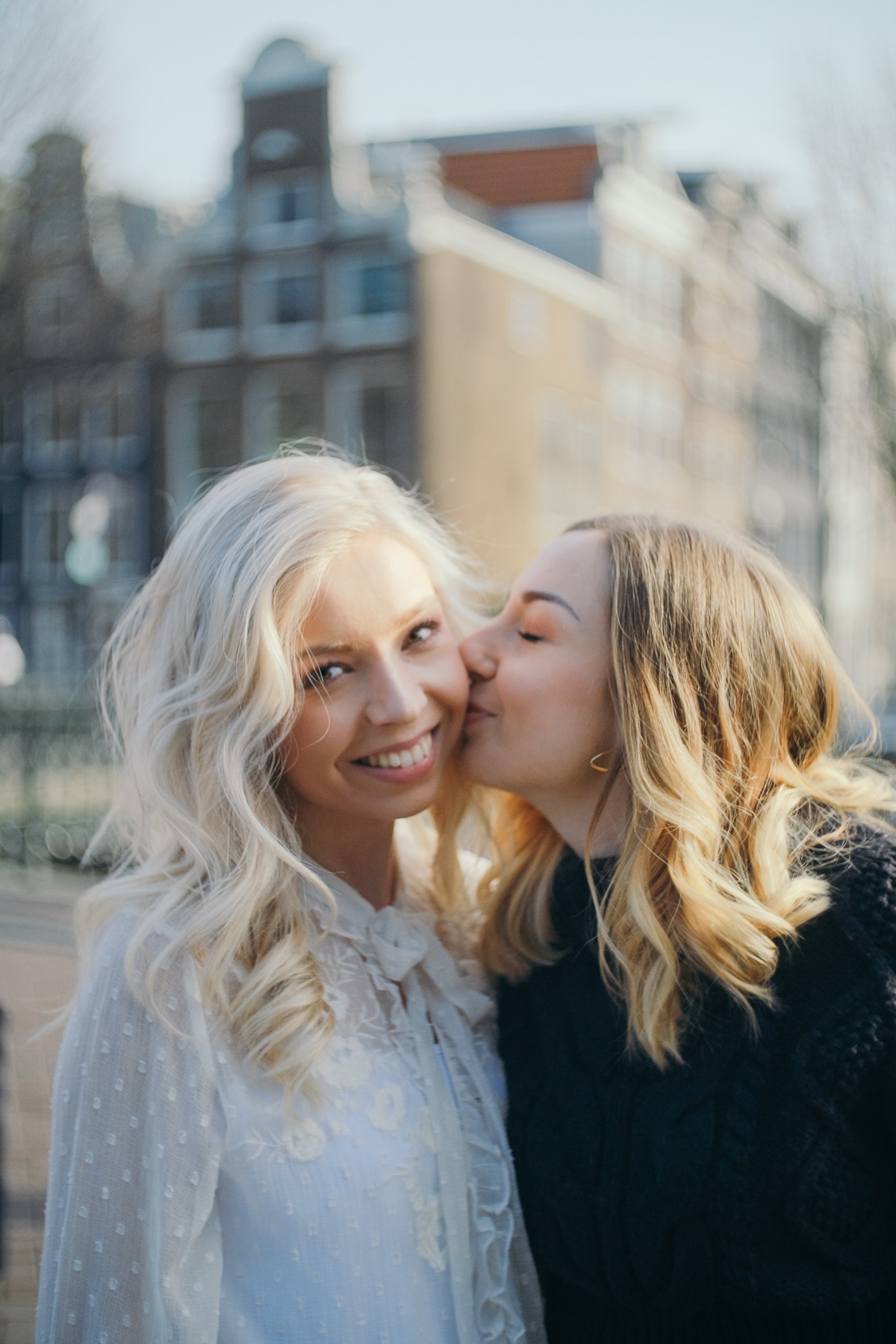 Friend kissing the blonde bride-to-be on the cheek, sunlit gabled houses soft in the background