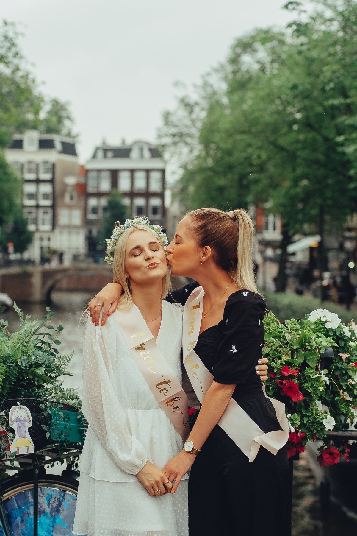Friend kissing the bride-to-be on the cheek on a canal bridge, her eyes closed under a crown of white flowers