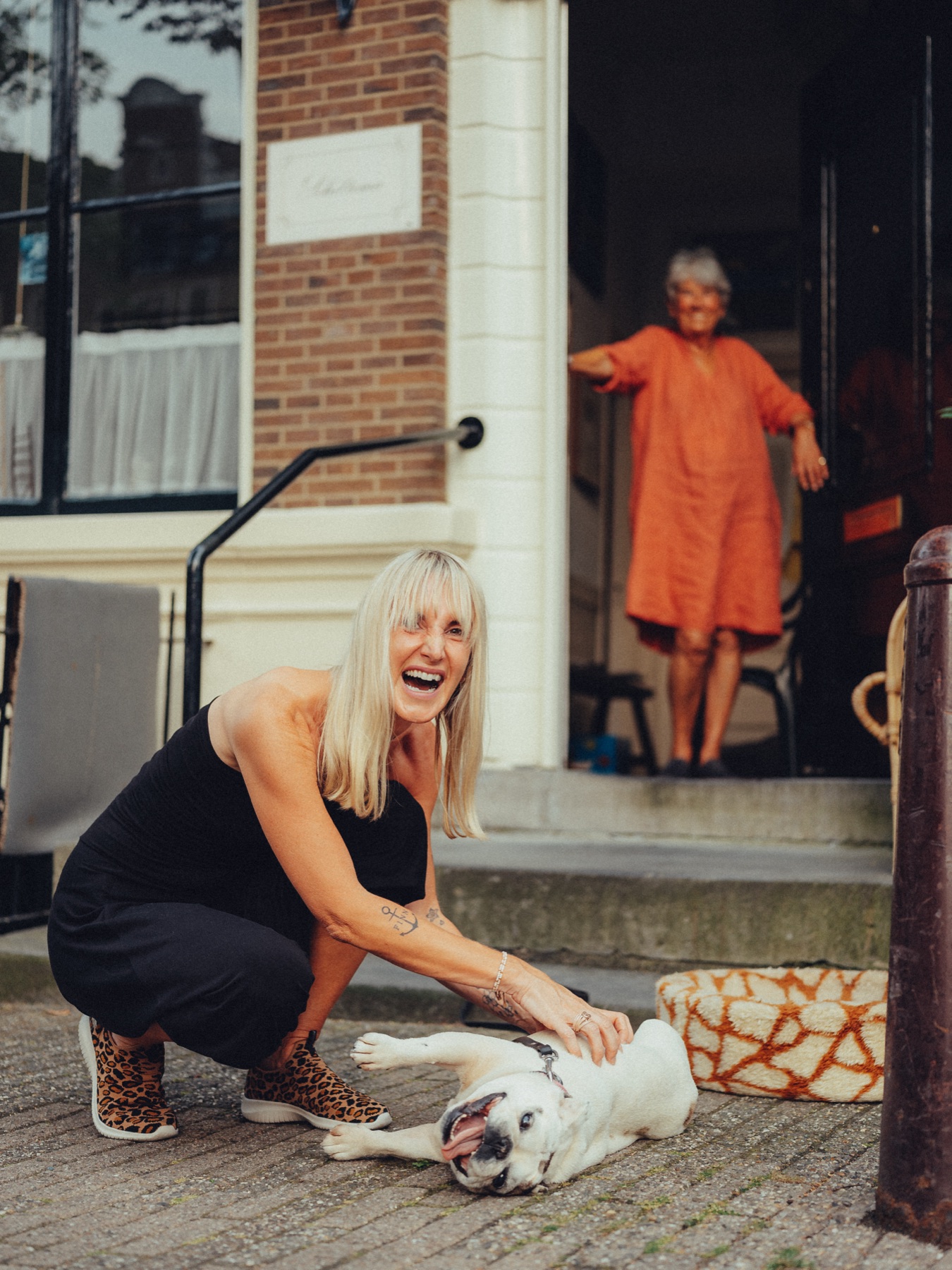 Friend in a black jumpsuit crouching to pet a white French bulldog rolling on its back, a woman in orange in the doorway