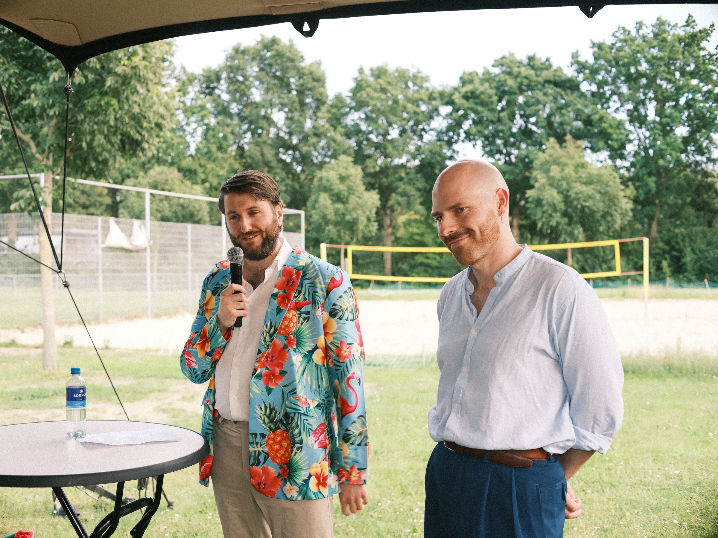 Friend in a jacket printed with tropical flowers and flamingos speaking into a microphone, a bald friend smiling beside him